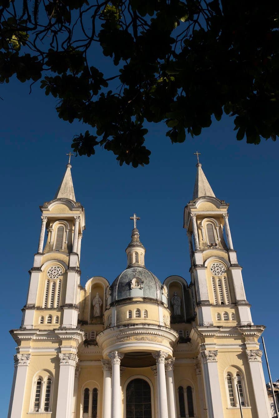 Ilhéus stellt sich gerne vor, laut, weiß und mitten in der Sonne. Froschperspektive auf eine blassgelbе und weiße neoklassizistische Kirche mit zwei Glockentürmen, einer zentralen Kuppel mit Kreuz und der Inschrift 'In Honorem Sancti Sebastiani' an der Fassade, eingerahmt von dunklen Ästen vor tiefblauem Himmel in Ilhéus, Bahia.