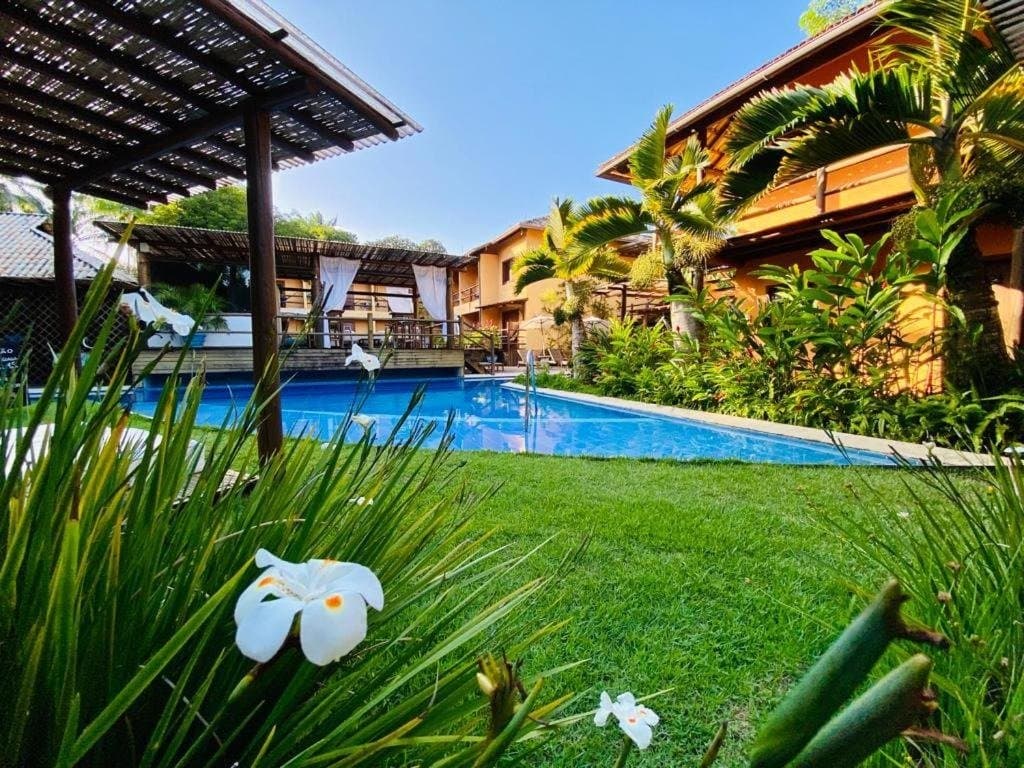 Hart geradelt, weich gelandet, so soll das sein. Üppiger Tropengarten mit weißen Blüten im Vordergrund, blauem Pool und einem warmtonigen zweistöckigen Gästehaus unter klarem Himmel in Itacaré