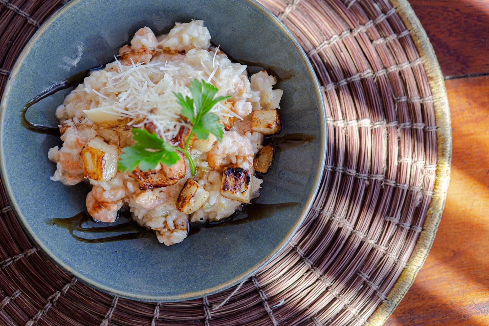 Nach dem Sattel kommt der Löffel. So soll das sein. Eine Keramikschale mit cremigem Garnelen-Risotto, angekohlten Wurzelgemüsestücken, geriebenem Käse, einem frischen Kräuterblatt und Balsamico-Träufeln, serviert auf einem geflochtenen Rattanuntersetzer auf einem Holztisch