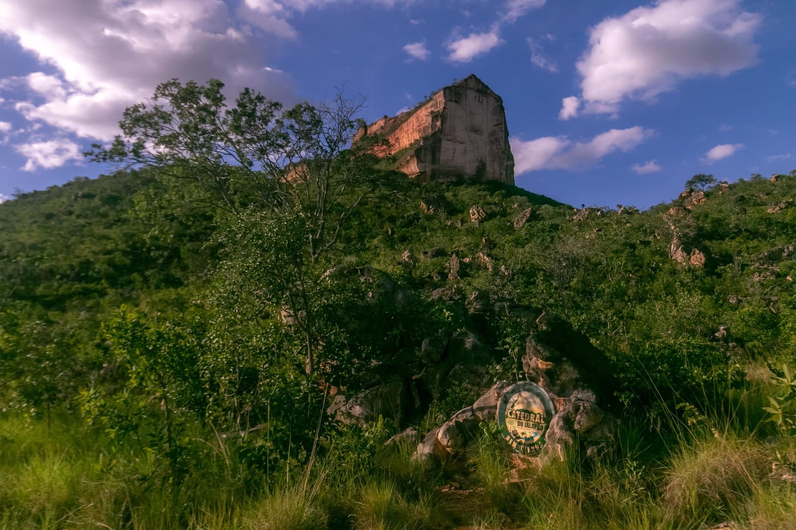 A placa diz onde você está; a pedra explica por que você veio. Photo: Alexandre Socci Vista em contra-plongée da Catedral do Jalapão, uma imponente formação rochosa de arenito que se ergue acima de uma encosta coberta de vegetação do Cerrado, com uma placa de trilha visível entre as pedras em primeiro plano e céu azul com nuvens esparsas