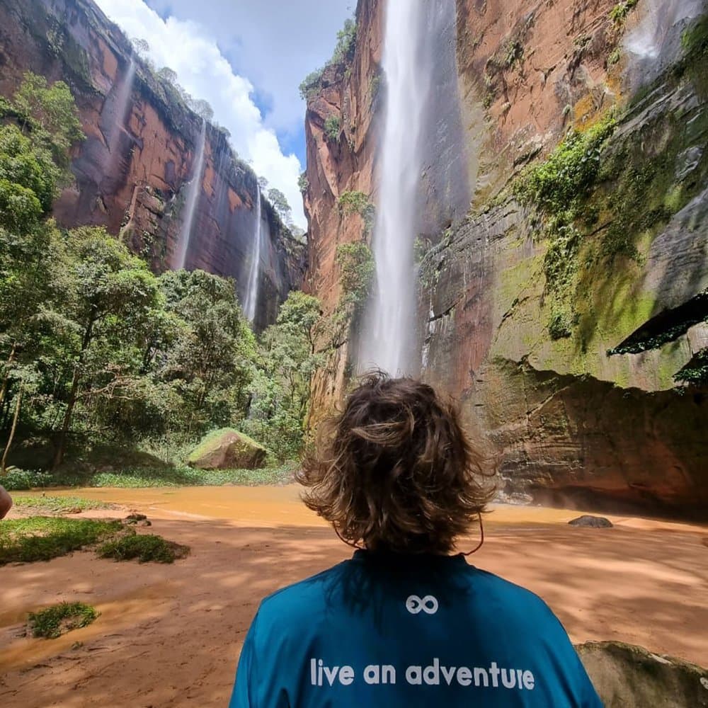 Cinco cachoeiras, uma vista só sua, e o almoço ainda por vir. Um viajante com camiseta azul-petróleo contempla múltiplas cachoeiras caindo por altas paredes de arenito vermelho até uma lagoa de cor ocre durante uma aventura em Jalapão