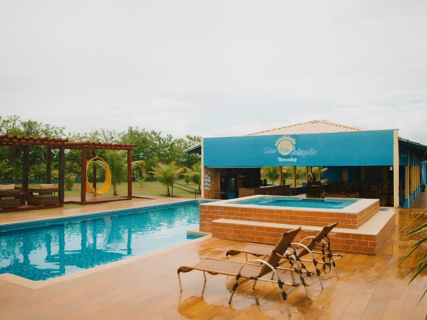 Trilha boa, mas a piscina no fim do dia tem seu charme. Piscina externa e hidromassagem elevada da Pousada São Félix do Tocantins, com espreguiçadeiras de vime em deck de cerâmica, uma cadeira suspensa amarela sob um pergolado e a fachada azul da pousada ao fundo — base confortável para uma aventura no Jalapão