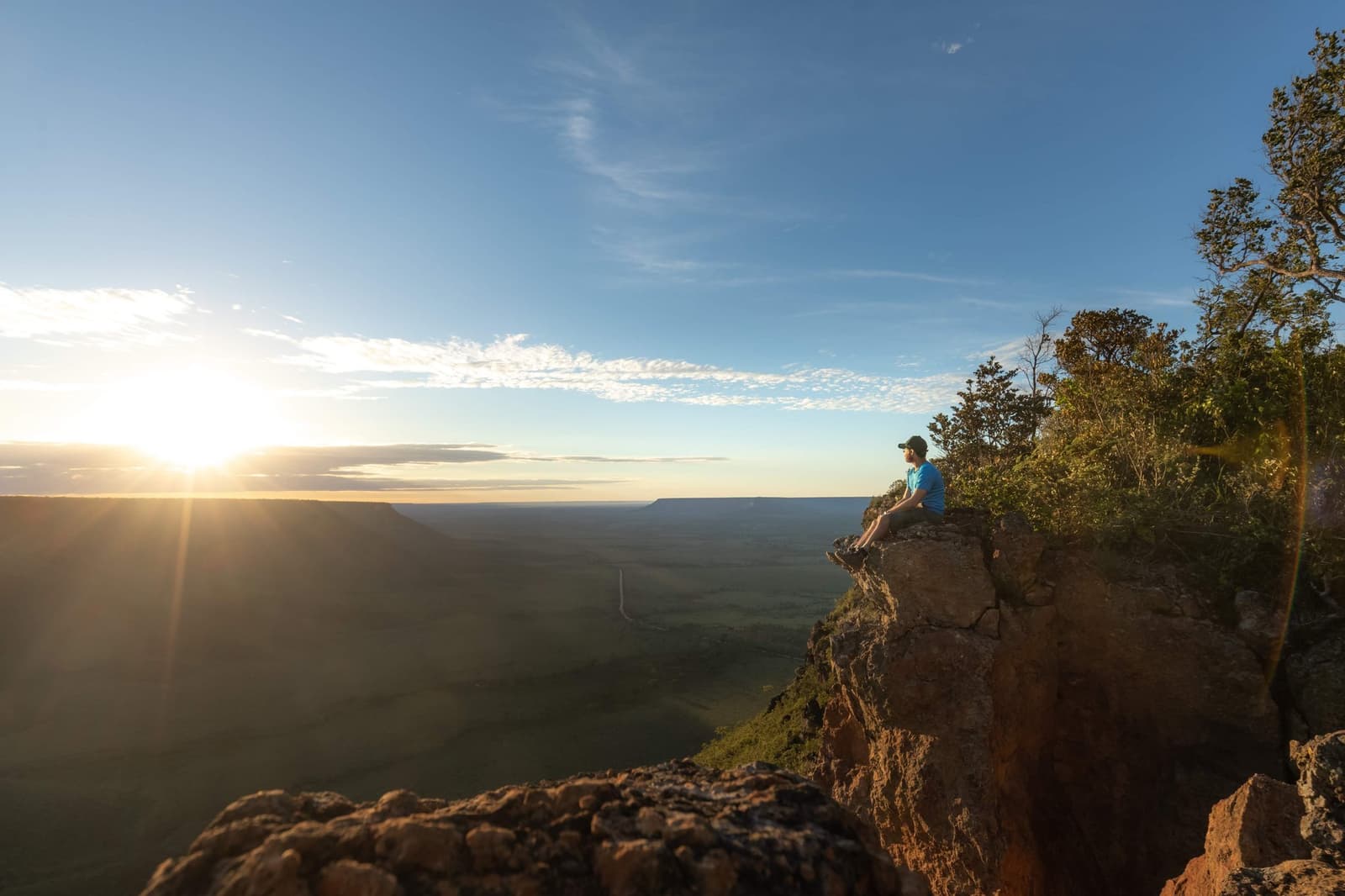 Antes do sol, já no alto. Vale cada degrau da subida. Photo: Alexandre Socci Um viajante sentado na beira de um penhasco ao amanhecer durante uma aventura no Jalapão, contemplando um vasto planalto do Cerrado com luz dourada se espalhando pelo horizonte