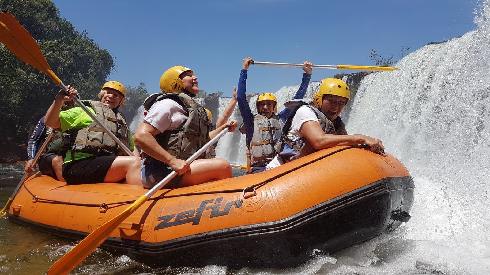 Paddel hoch, Gesichter klatschnass, der Novo River entscheidet, wer hier das Sagen hat. Fünf lachende Personen mit gelben Helmen und Schwimmwesten paddeln auf einem orangefarbenen Schlauchboot an einem rauschenden Wasserfall vorbei beim Jalapão-Trekking und Rafting