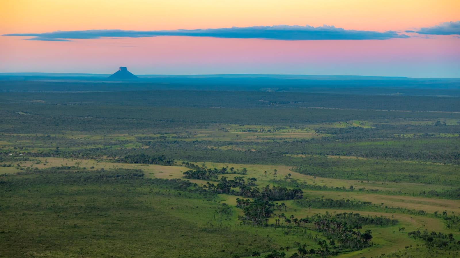 Vier Stunden ab Palmas, und plötzlich ist die Welt so groß wie das hier. Photo: Alberto Andrich Luftpanorama einer weiten Cerrado-Savanne in der Abenddämmerung auf einem Jalapão-Trip, mit einem einzelnen Tafelberg als Silhouette vor einem rosa-orangefarbenen Himmel am Horizont