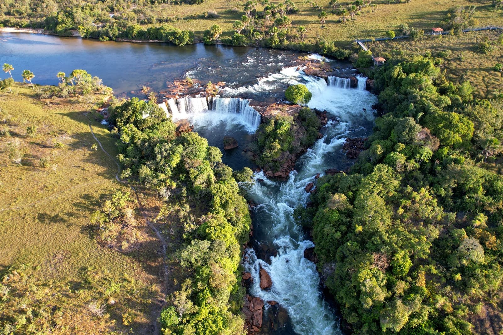 Der Fluss kennt nur eine Richtung, und er meint es ernst. Photo: Wirestock Luftaufnahme eines breiten mehrstufigen Wasserfalls im Jalapão, wo ein Fluss über eisenhaltige Felsvorsprünge in eine schäumende Schlucht stürzt, flankiert von dichtem Galeriewald und offenem Grasland