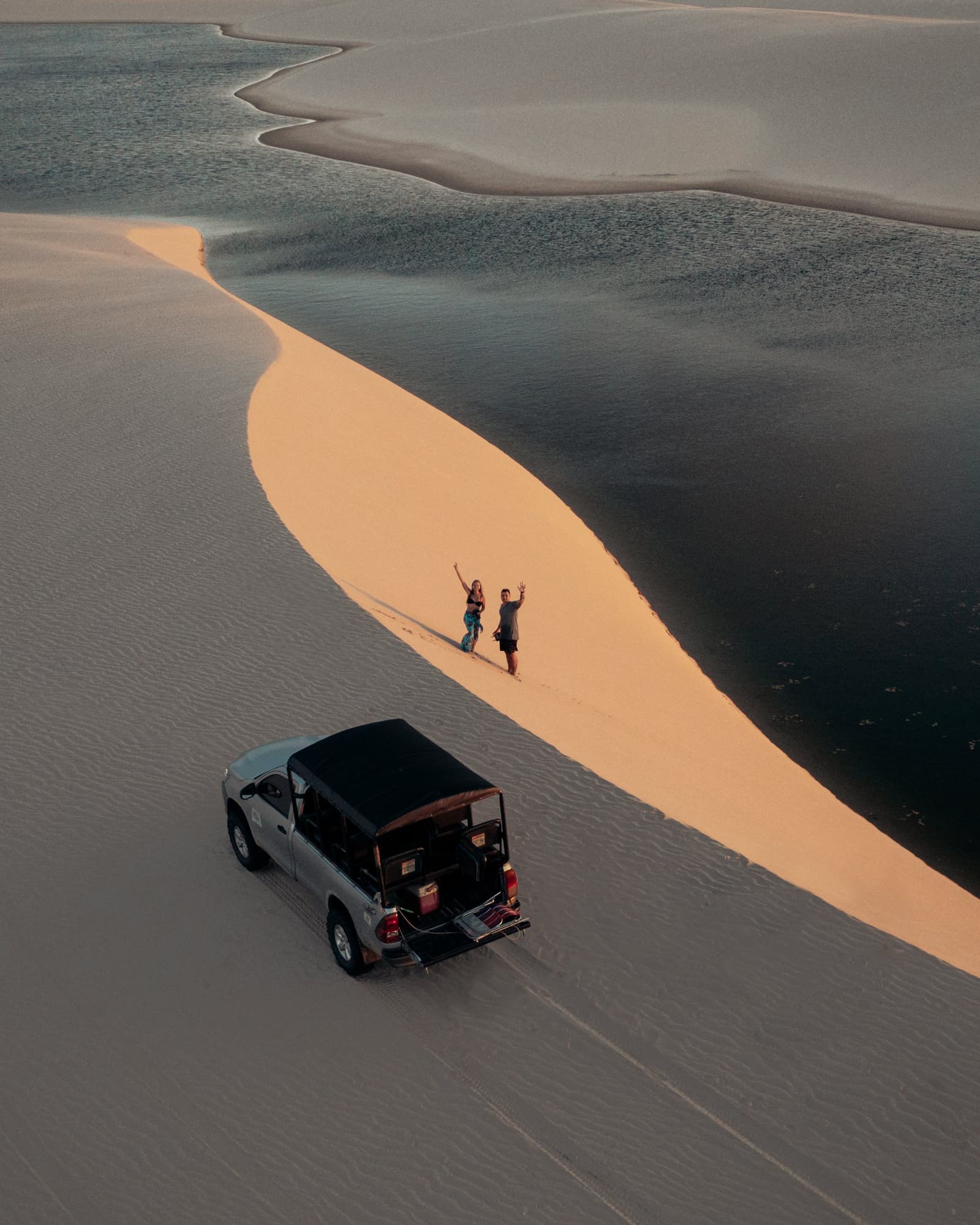 Kein Netz, kein Problem, nur Sand, Wasser und diese Weite. Luftaufnahme zweier Reisender, die bei Sonnenuntergang auf einem goldenen Dünenkamm neben einer dunklen Lagune winken, mit einem 4x4-Fahrzeug darunter — Teil eines Lençóis Maranhenses Reiseprogramms