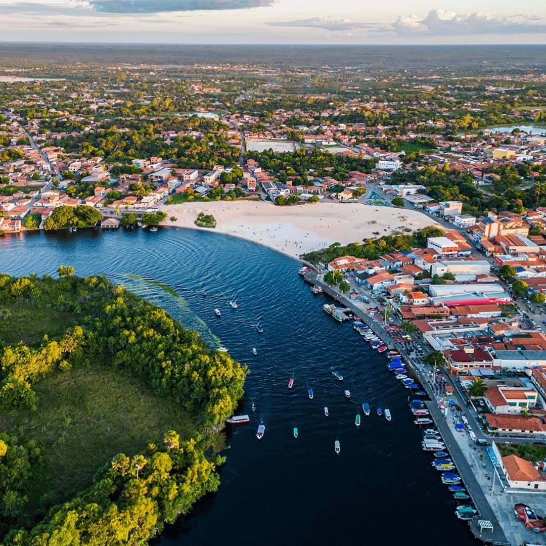 Eine Stadt, die sich eng ans Flussufer schmiegt, und das aus gutem Grund. Luftaufnahme von Barreirinhas mit dem Fluss Preguiças, einem weißen Sandstrand, bunten Booten am Ufer und der Stadt inmitten grüner Vegetation – idealer Ausgangspunkt für das vollständige Lençóis-Maranhenses-Reiseprogramm