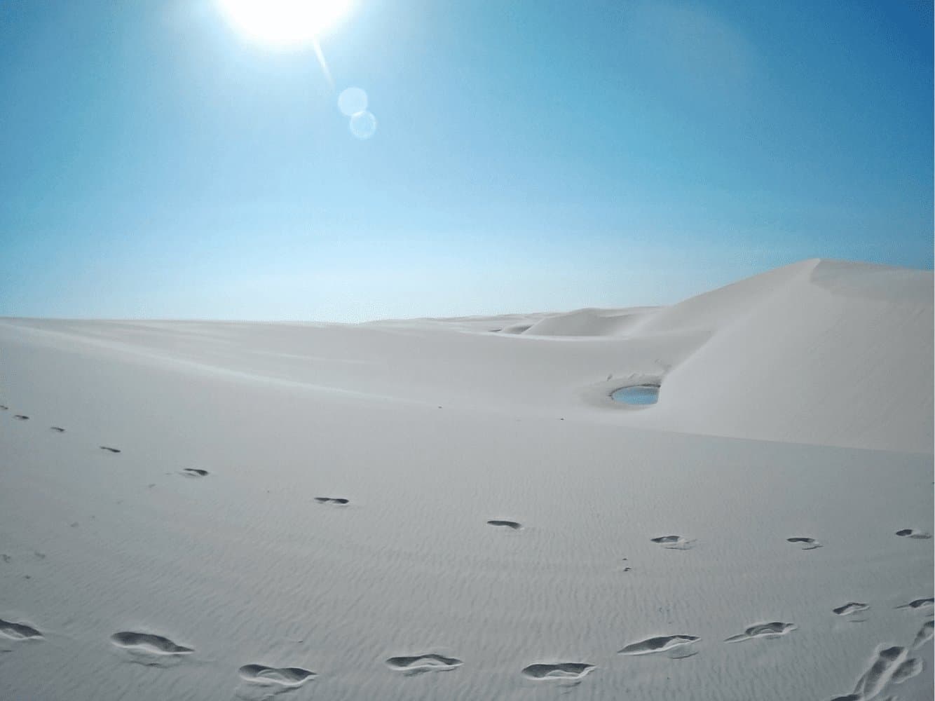 Jemand ist hier entlanggegangen, hat Wasser gefunden und seinen Augen kaum getraut. Fußabdrücke im weißen Sand der Lençóis Maranhenses führen auf eine kleine blaue Lagune zwischen sanften Dünenrücken zu, unter gleißender Mittagssonne