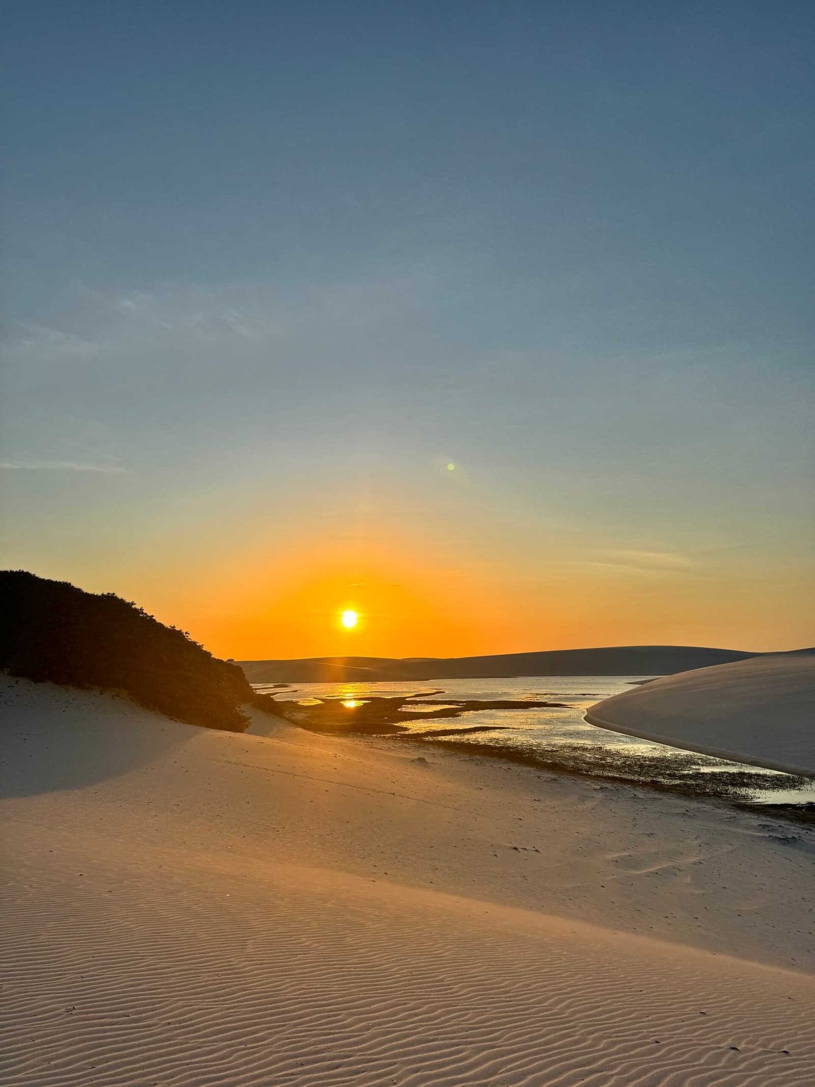 Das letzte Licht des Tages, festgehalten zwischen Sand und Wasser. Goldener Sonnenuntergang über einem Gezeitenkanal zwischen weißen Sanddünen in Lençóis Maranhenses, mit orangefarbenem Lichtreflex auf dem flachen Wasser