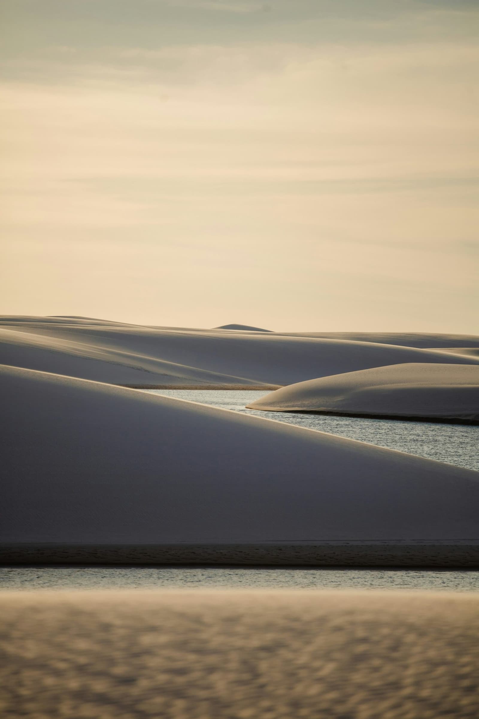 Sand und Wasser, die sich jede Saison neu erfinden. Geschichtete weiße Sanddünen werfen tiefe Schatten auf eine ruhige Süßwasserlagune im Lençóis Maranhenses, unter einem weichen, warmen Himmel