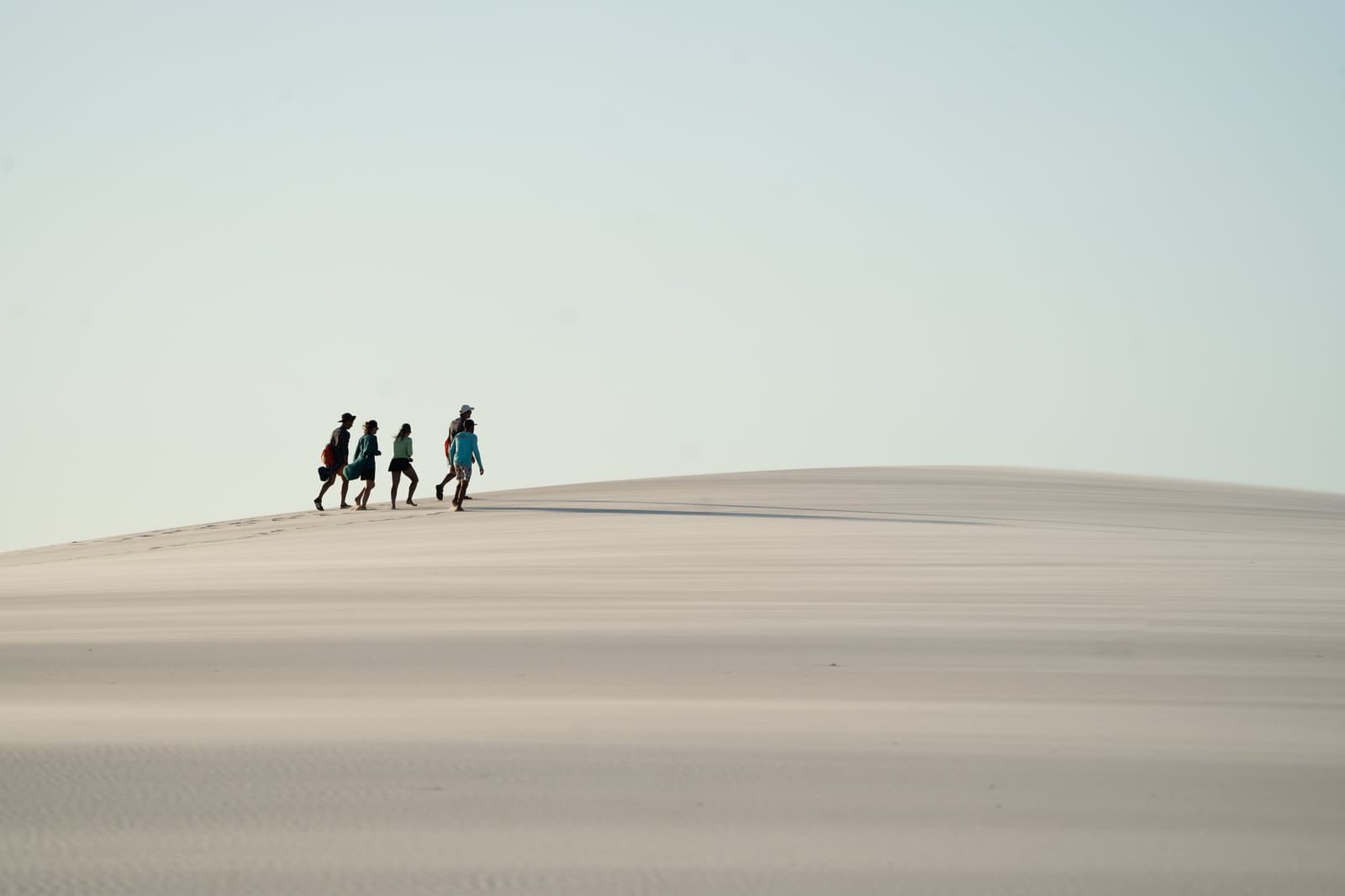Auf dem Dünenkamm wird klar, wie klein wir wirklich sind. Vier Wanderer schreiten entlang des geschwungenen Kamms einer riesigen weißen Sanddüne bei einer Lençóis-Maranhenses-Wanderung unter hellblauem Himmel