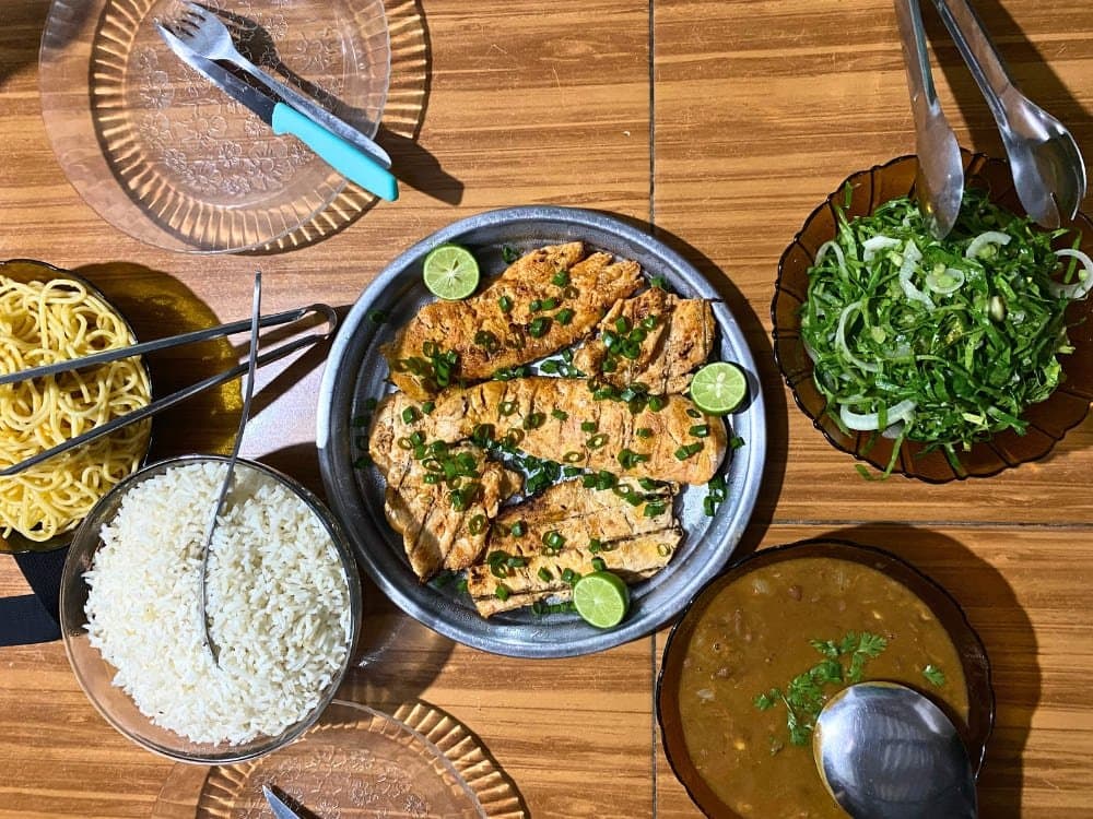 So sieht Mittagspause aus, wenn das Maranhão kocht. Vogelperspektive auf ein selbst gekochtes Mittagessen beim Lençóis-Maranhenses-Trekking: gegrillte Fischfilets mit Frühlingszwiebeln und Limettenscheiben auf einem Metallteller, dazu Schüsseln mit weißem Reis, Nudeln, grünem Salat und einer braunen Bohnenbrühe