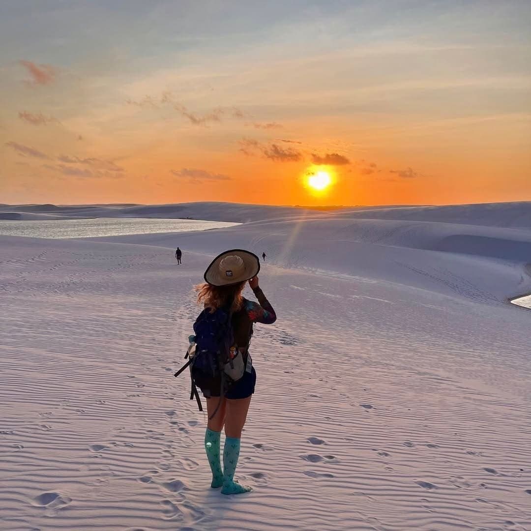 Sonne versinkt, Hut kaum zu halten, manchmal braucht es genau diesen Moment. Eine Wanderin mit breitkrempigem Hut und Rucksack steht in türkisfarbenen Socken auf weißen Sanddünen beim Lençóis-Maranhenses-Trekking und beobachtet einen orangefarbenen Sonnenuntergang über fernen Lagunen