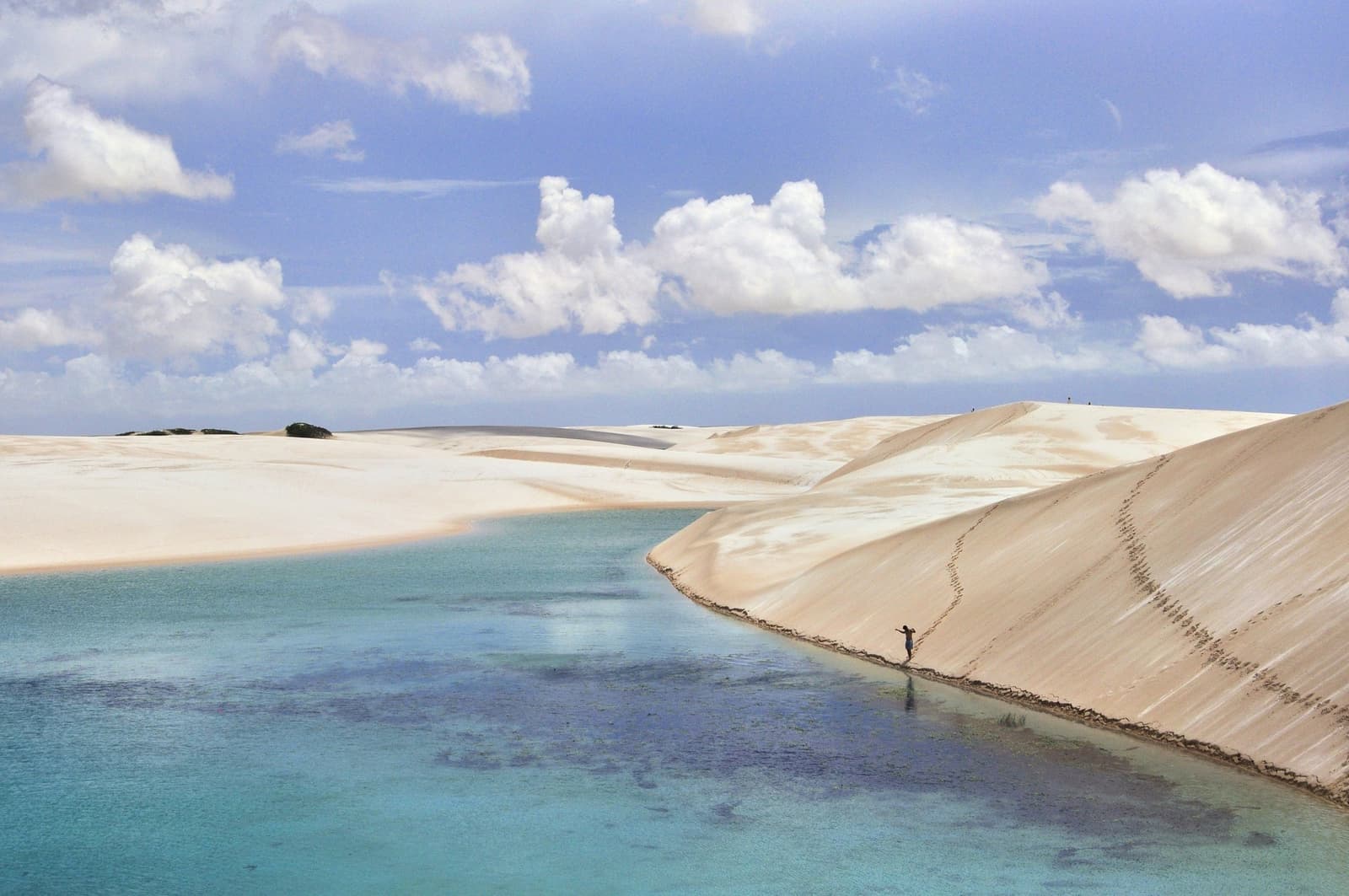 Ein Mensch, eine Lagune, und Dünen, die alles in Relation setzen. Eine einzelne Person wandert am Rand einer weißen Sanddüne entlang einer türkisfarbenen Lagune in den Lençóis Maranhenses, mit Wolken, die sich im stillen Wasser spiegeln
