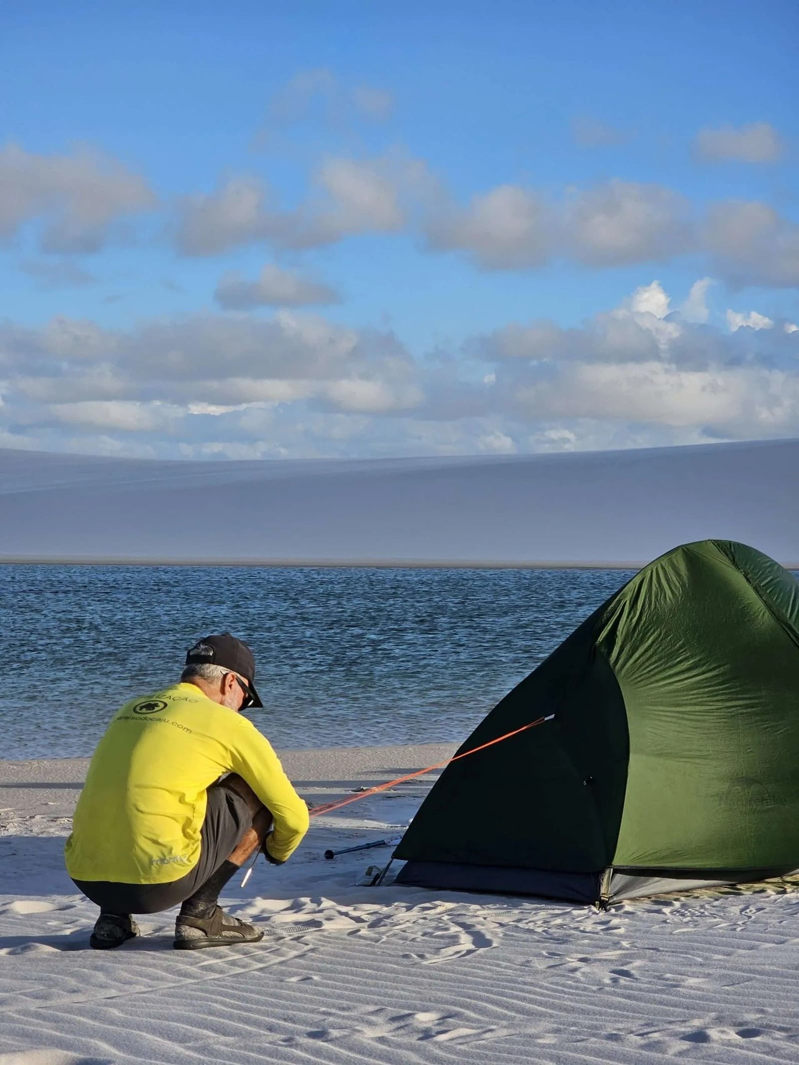Le camp est posé, les dunes peuvent attendre le lever du soleil. Un guide en t-shirt jaune s'accroupit pour planter un piquet de tente verte sur le sable blanc au bord d'une lagune bleue calme, lors d'un trek dans les Lençóis Maranhenses, avec les dunes visibles au loin