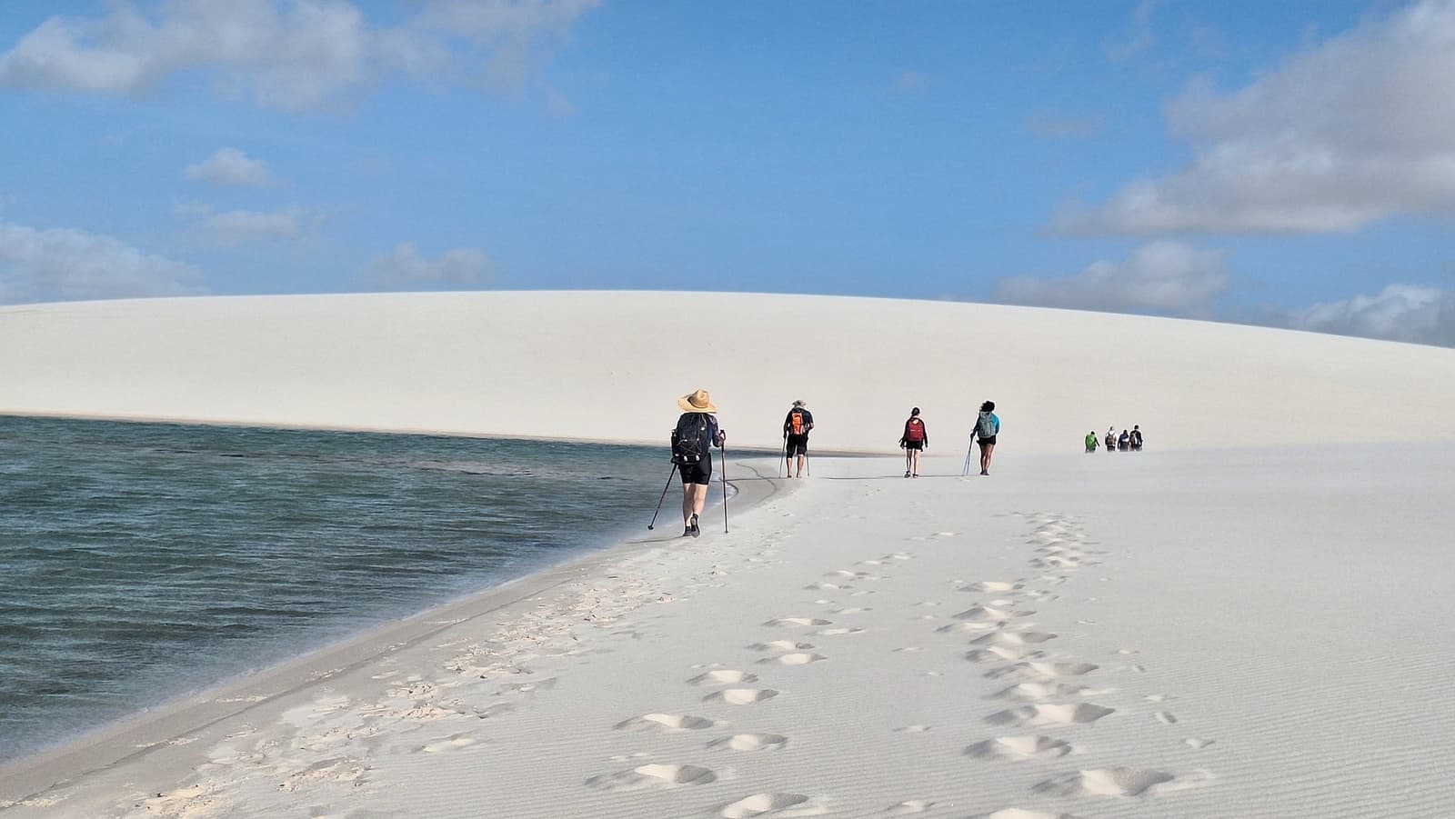 Un lagon à gauche, une dune à droite, et des pas qui s'enfoncent dans le sable. Un groupe de randonneurs équipés de bâtons de marche et de sacs à dos avance sur une mince bande de sable blanc entre une lagune aux eaux sombres et une immense dune blanche lors d'un trek dans les Lençóis Maranhenses