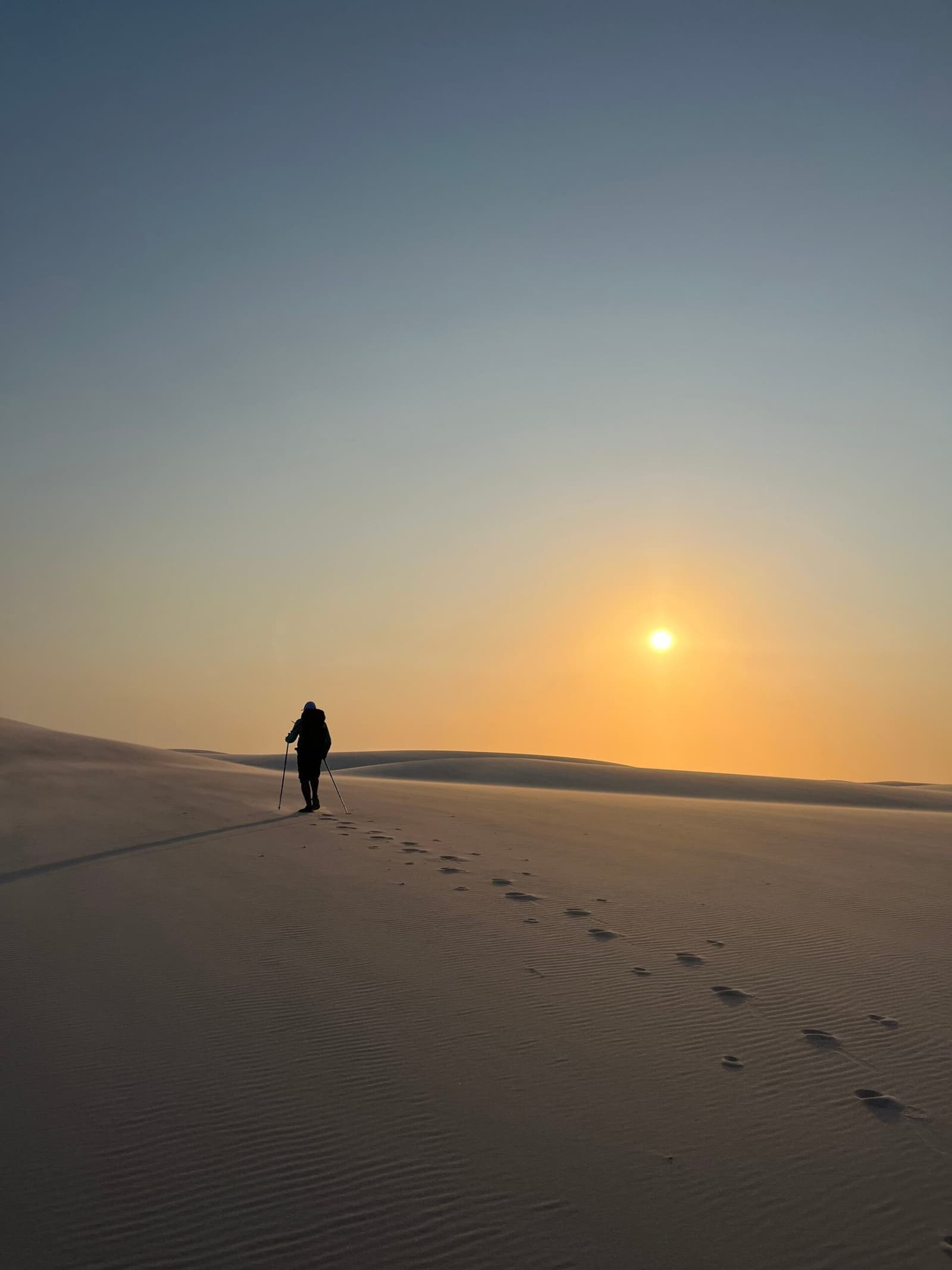 Des empreintes dans le sable, et tout le reste peut attendre. Un randonneur solitaire avec des bâtons de marche gravit une vaste dune de sable blanc lors d'un trek dans les Lençóis Maranhenses, laissant une longue trace d'empreintes sous un soleil levant doré