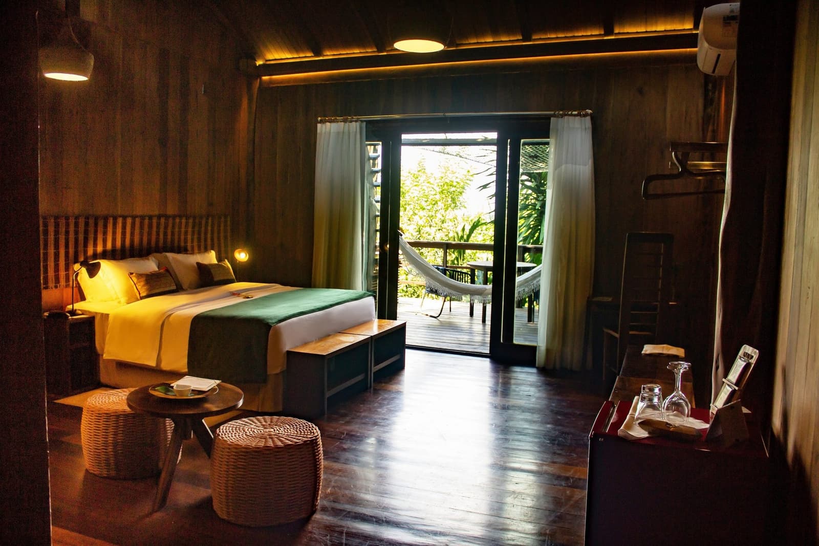 Holzwände, Hängematte, Dschungelblick, mehr braucht man nicht. Gemütlich beleuchtetes Holzkabinenzimmer im Mirante do Gavião, mit Doppelbett in Gelb und Grün, Korbhockern und offenen Glastüren zu einem privaten Balkon mit Hängematte und Blick in den Regenwald