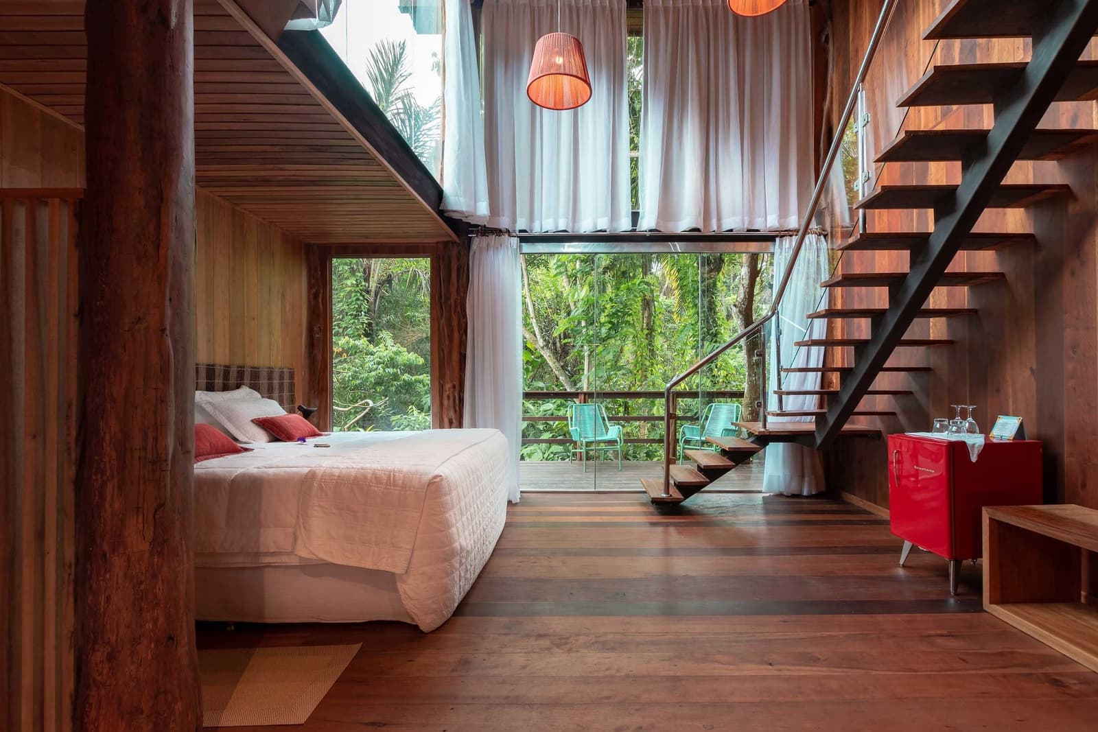 Holz, Grün und ein Bett, das keine weiteren Pläne verlangt. Loft-Suite im Mirante do Gavião Amazon Lodge mit warmen Holzwänden, Doppelbett mit roten Akzentkissen, freischwebender Stahltreppe und bodentiefen Glasfronten, die auf einen privaten Balkon mit Blick in den Regenwald führen