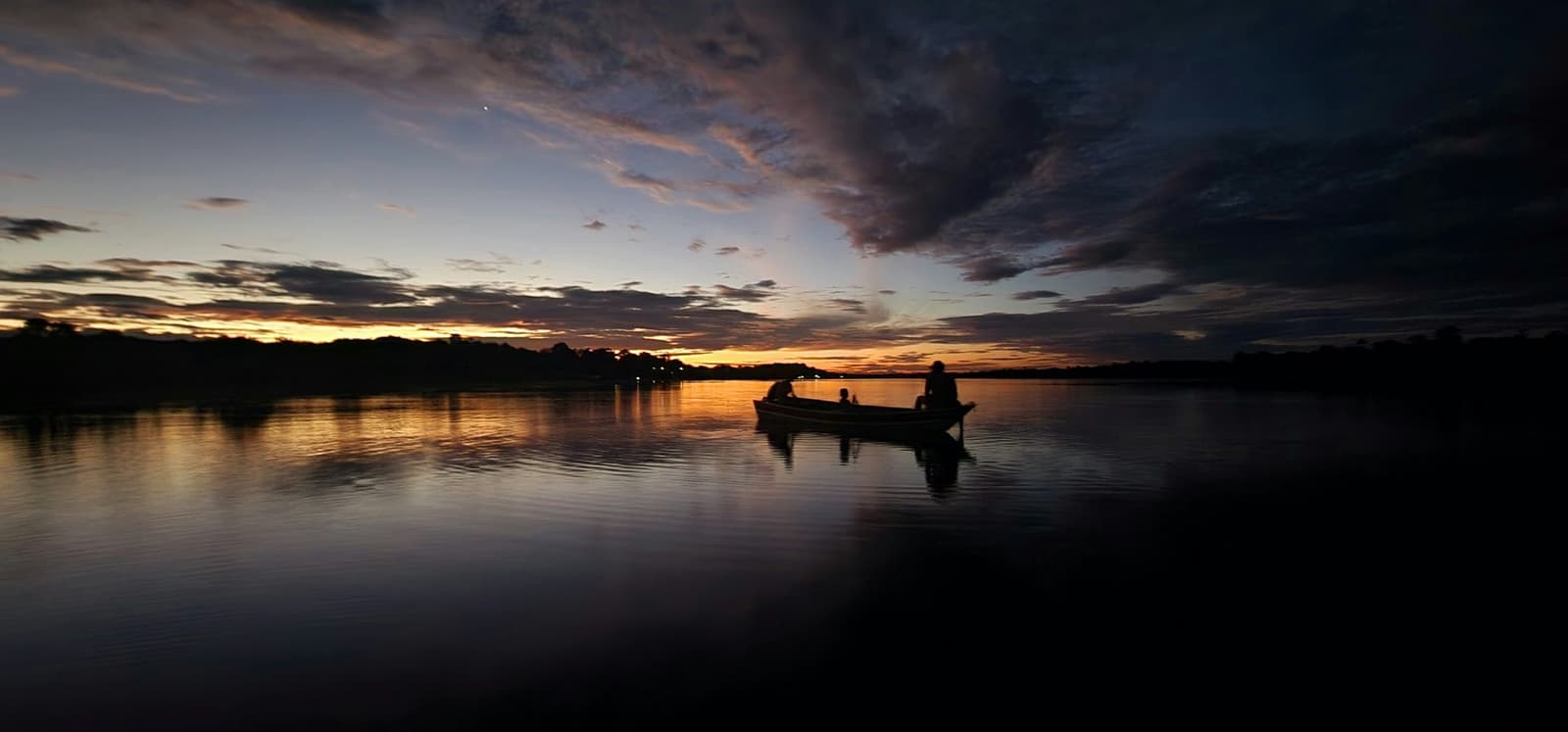 Der Fluss schluckt das letzte Licht, und niemand will zurück. Silhouette von drei Personen in einem kleinen Holzboot auf spiegelglattem Wasser in der Abenddämmerung, der Sonnenuntergang am Mirante do Gavião taucht den Horizont in Gold und Violett