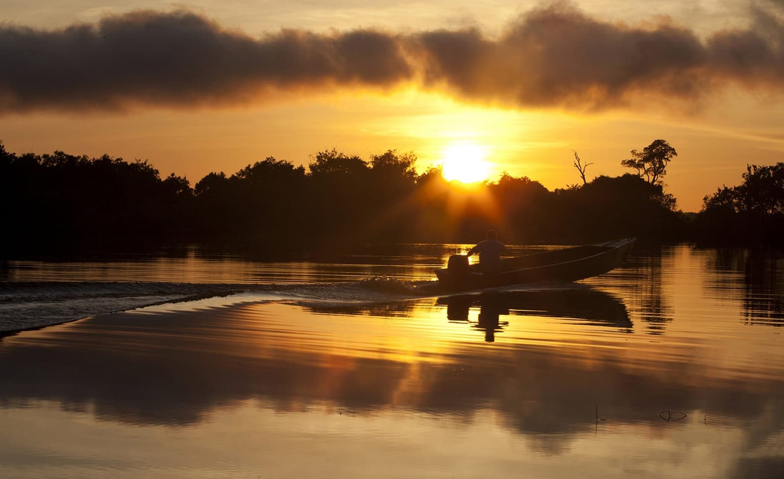 Der Fluss hat seinen eigenen Rhythmus, und dieser Moment gehört zu den schönsten. Eine Silhouette steuert ein Motorboot über einen spiegelglatten Fluss im goldenen Sonnenuntergang beim Mirante do Gavião, die Sonne bricht durch Wolken über einer dunklen Baumsilhouette