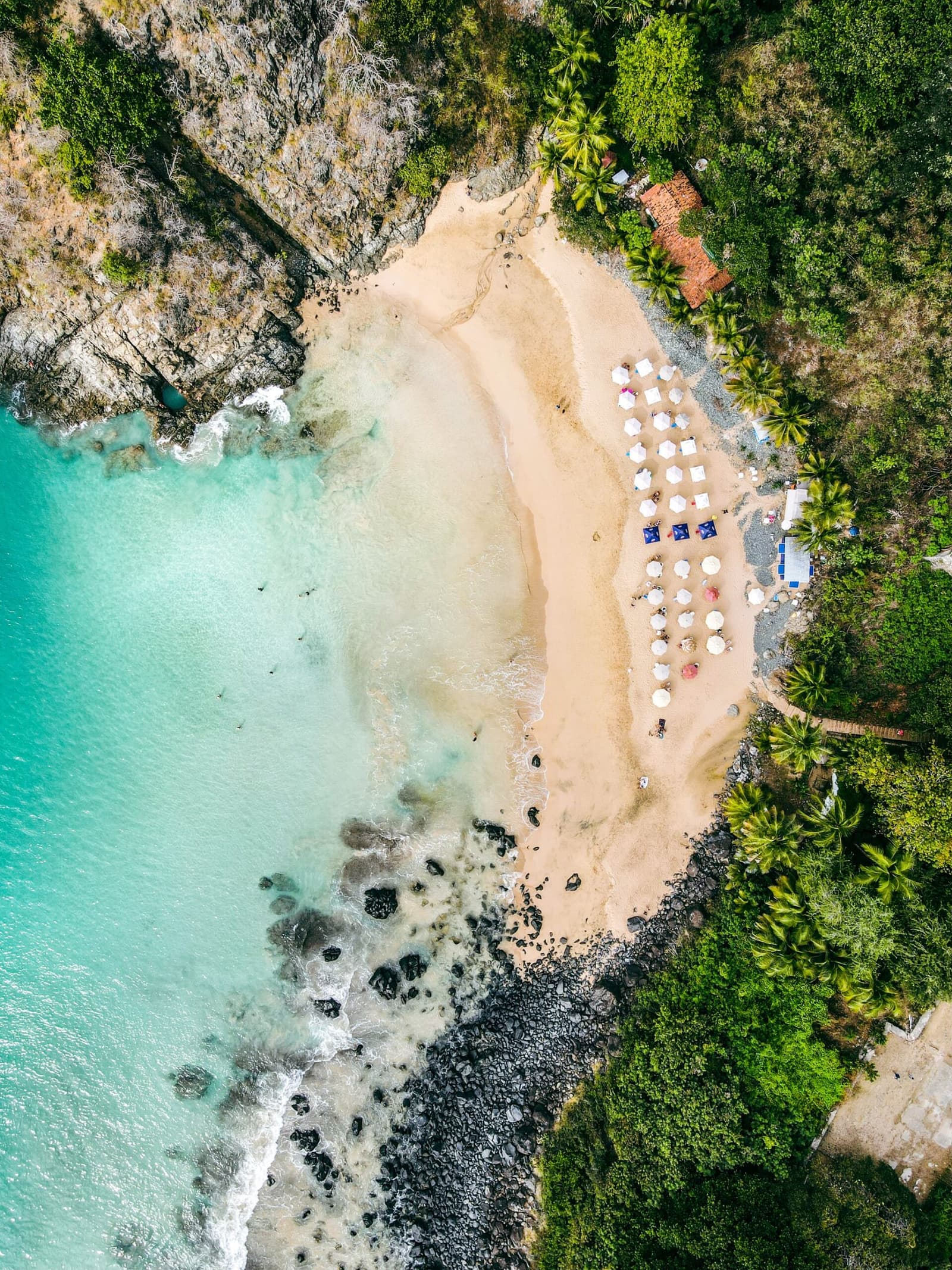 Genau so sieht es aus, bevor man zum ersten Mal abtaucht. Drohnenaufnahme einer kleinen Sandbucht in Fernando de Noronha, eingebettet zwischen Felsen und üppiger Vegetation, mit Reihen weißer Sonnenschirme und türkisfarbenem Wasser — ein geschützter Ort für die ersten Strandtauchgänge beim Open Water Tauchkurs