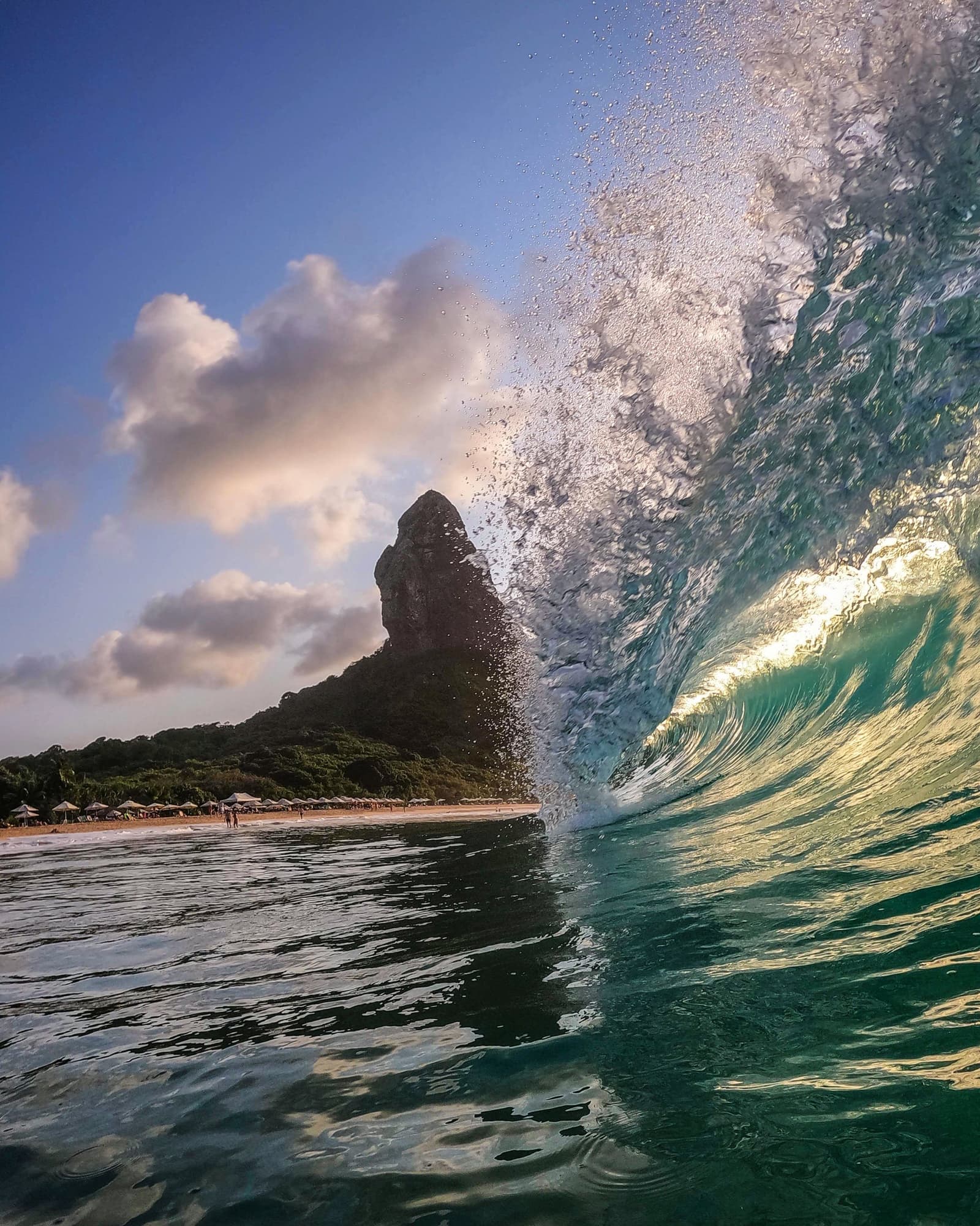 Das Meer hier hat eine klare Meinung, und es macht sie lautstark deutlich. Eine türkisblaue Welle bricht schäumend auf, während im Hintergrund der markante Felsturm Morro do Pico und ein palmengesäumter Strand in Fernando de Noronha unter blauem Himmel zu sehen sind.