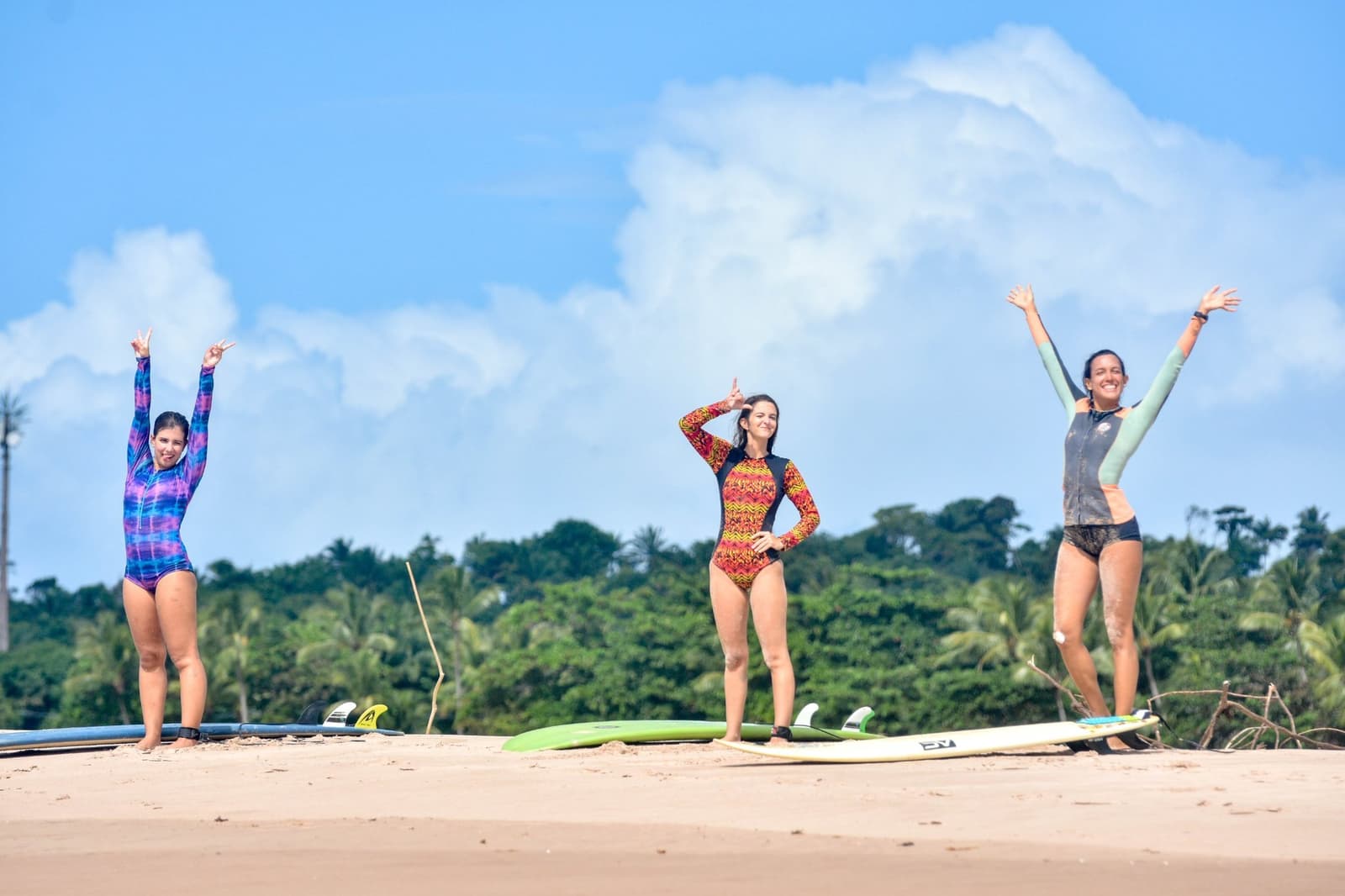 Sorriso no rosto, prancha no chão, e a aula ainda nem terminou. Photo: Picasa Três mulheres com lycras coloridas comemoram na areia com suas pranchas durante aulas de surf em Itacaré, com uma densa mata verde e céu azul ao fundo.