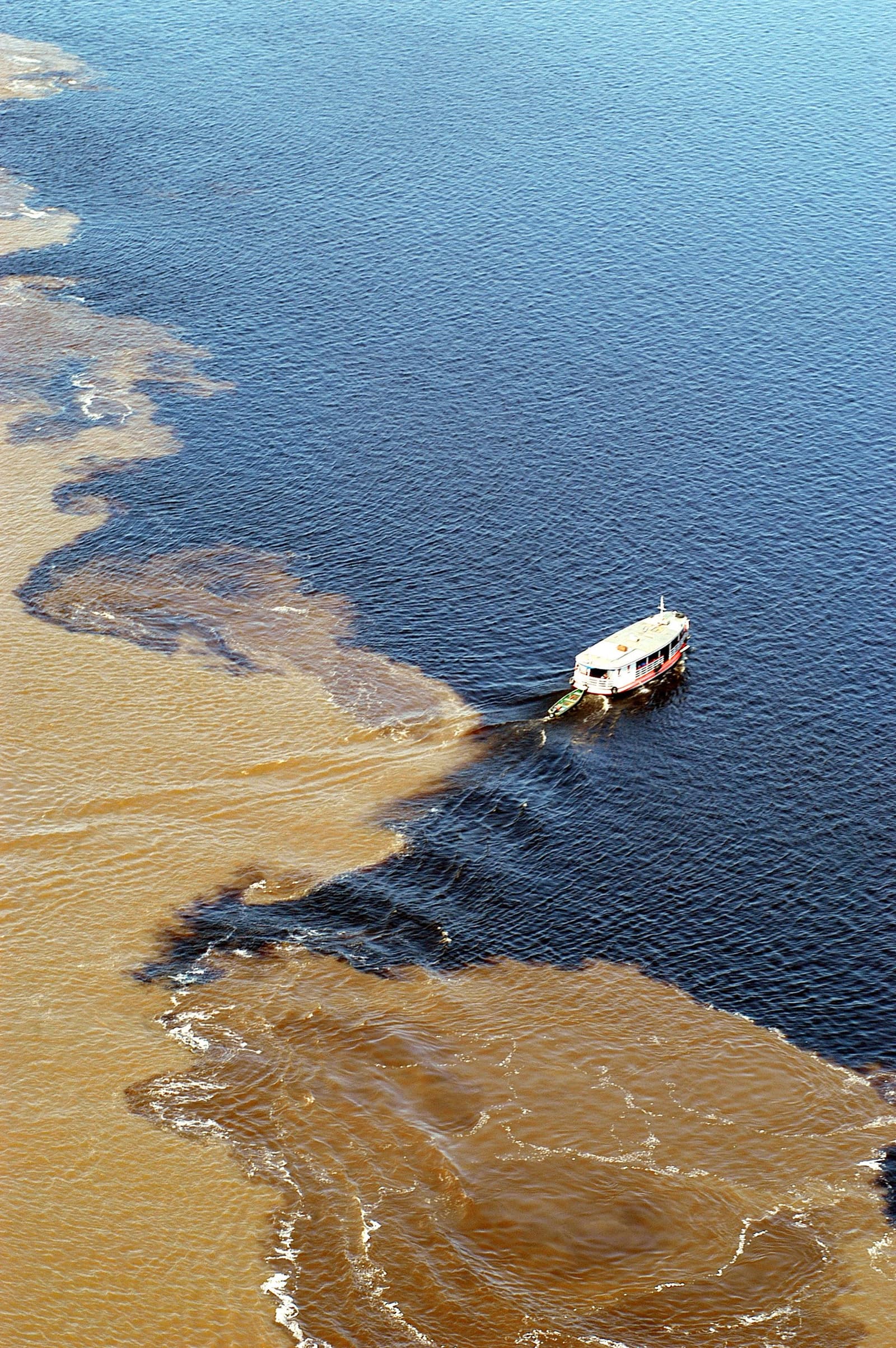 Zwei Flüsse, eine Grenze, kein Kompromiss. Luftaufnahme eines Flusskreuzfahrtschiffs beim Überqueren des Begegnungspunkts zweier Flüsse, wo dunkles Schwarzwasser und sandbraunes Sedimentwasser nebeneinander fließen, ohne sich zu vermischen
