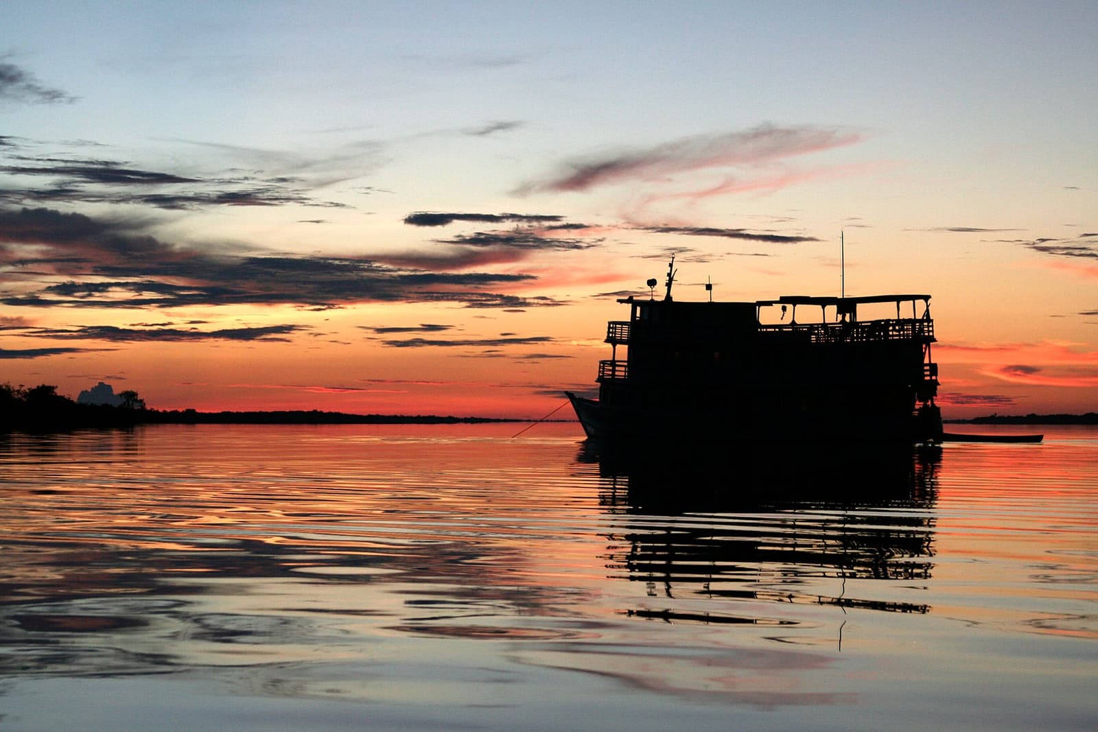 Das letzte Licht des Tages, und der Fluss gehört euch ganz allein. Ein mehrstöckiges Flussschiff als Silhouette vor einem leuchtendem orange-roten Sonnenuntergangshimmel, der sich im stillen dunklen Wasser spiegelt, auf der Tucano Amazon River Cruise