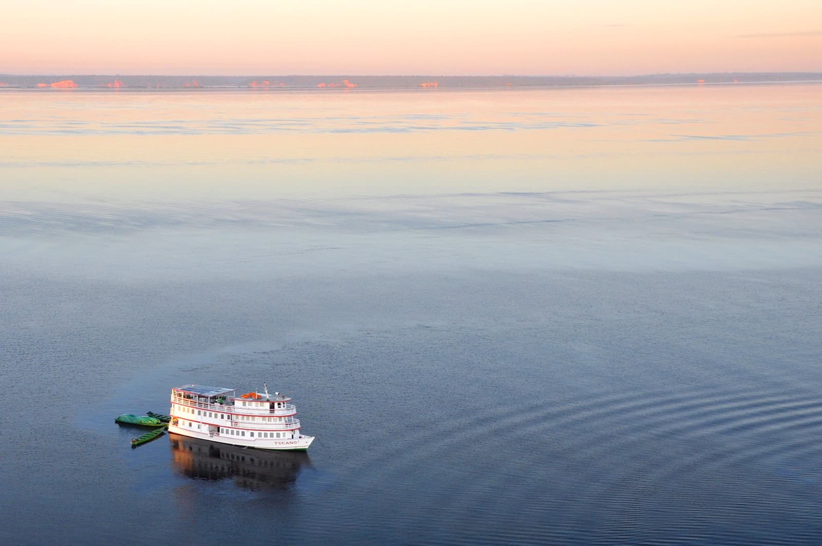 So viel Fluss, so wenig Eile, genau das ist der Plan. Das M/Y Tucano, ein weißes mehrstöckiges Schiff auf der Tucano Amazon River Cruise, gleitet in der Abenddämmerung über einen weiten, spiegelglatten Fluss unter einem zartrosa Himmel