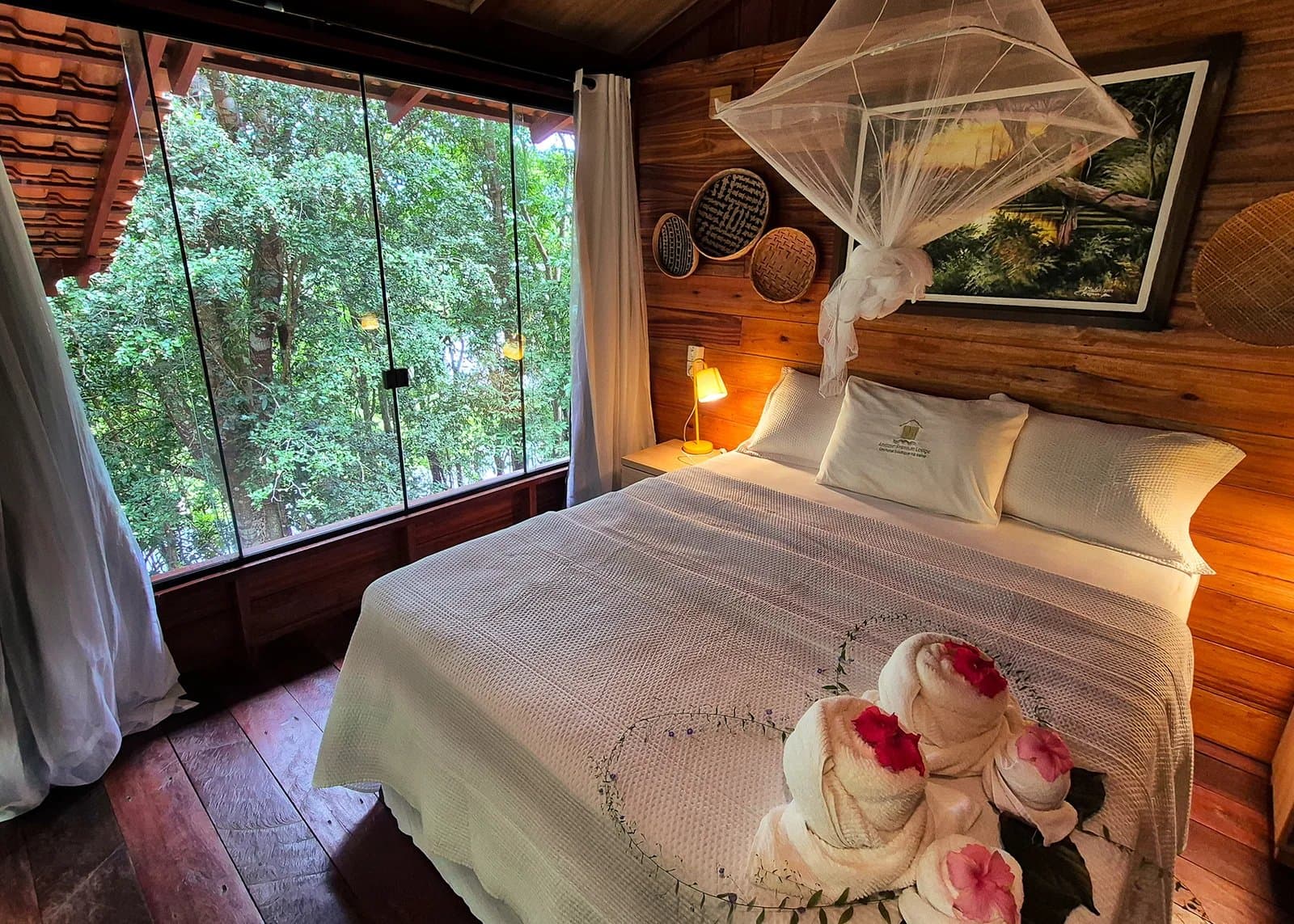 Handtücher als Kunstwerk, der Regenwald direkt vor dem Fenster. Gute Nacht. Gemütliches Schlafzimmer in einer Amazon-Boutique-Lodge mit Mostikonetz-Himmelbett, warmen Holzwänden, geflochtenen Körben als Deko, zu Blumen gefalteten Handtüchern und einem raumhohen Fenster mit Blick in den dichten Regenwald