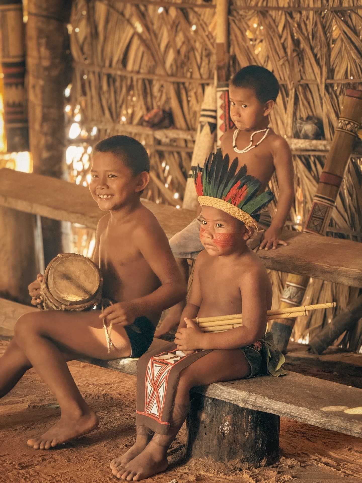 Musik, Farbe, Feder, diese drei haben ihren Auftritt längst verinnerlicht. Drei indigene Kinder in einer Reetdach-Maloca am Ariaú-Fluss während einer Amazonas-Kreuzfahrt in Brasilien – ein Junge lächelt und hält eine kleine Handtrommel, ein anderer trägt einen Federschmuck und rote Gesichtsbemalung, ein dritter schaut still von hinten zu