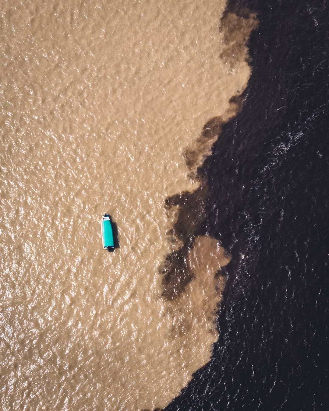 Zwei Flüsse, eine scharfe Linie, ein winziges Boot. Luftaufnahme eines Bootes mit türkisfarbenem Dach an der Begegnung der Gewässer, wo helles, sedimentreiches Wasser auf tiefschwarzes Wasser trifft