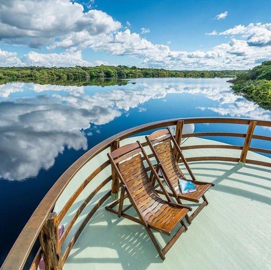 Zwei Stühle, ein Fluss, und ein Himmel, der oben und unten verwechselt. Zwei Holzliegestühle auf dem geschwungenen Bug eines Flussbootes bei einer Amazonas-Flusskreuzfahrt in Brasilien, mit Wolken, die sich auf dem spiegelglatten Schwarzwasserfluss reflektieren