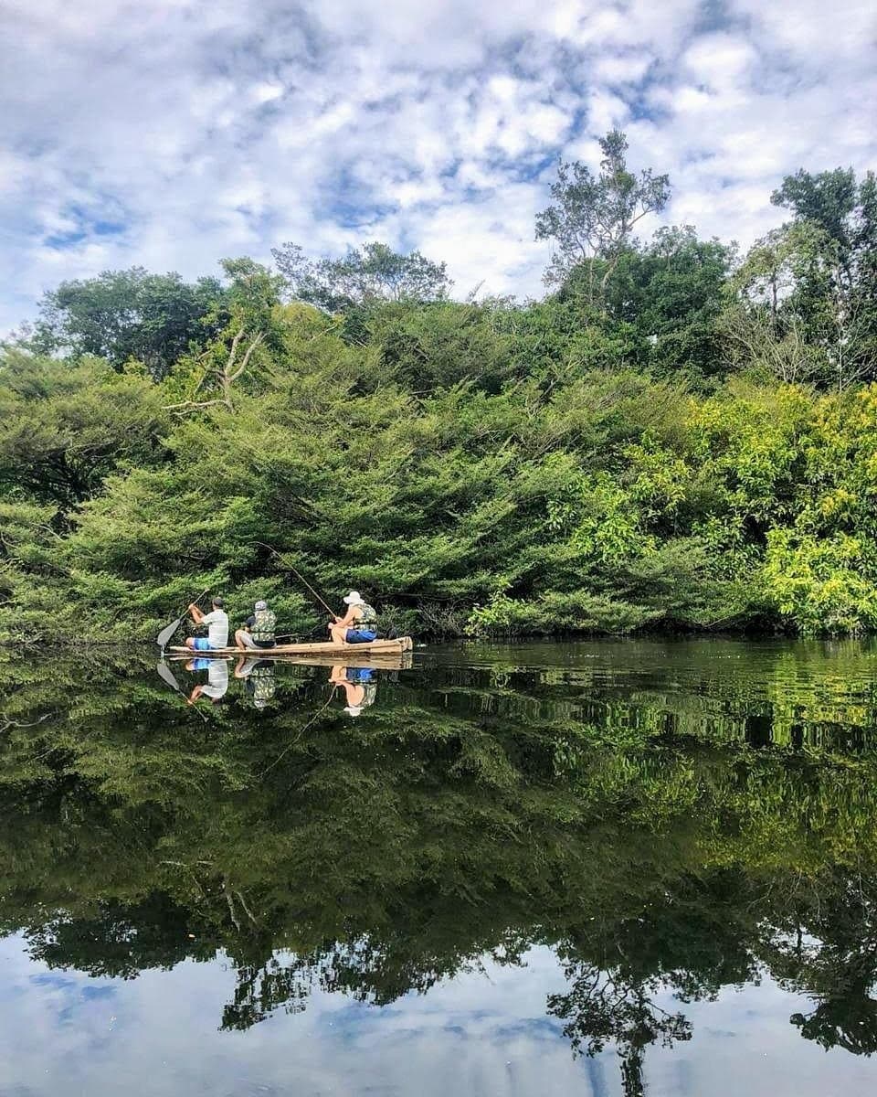Angeln, Stille, dichter Wald, und irgendwo da unten ein Piranha mit Meinung. Drei Gäste angeln von einem Holzkanu auf einem spiegelglatten Fluss, umgeben von dichtem tropischen Regenwald unter einem teils bewölkten Himmel