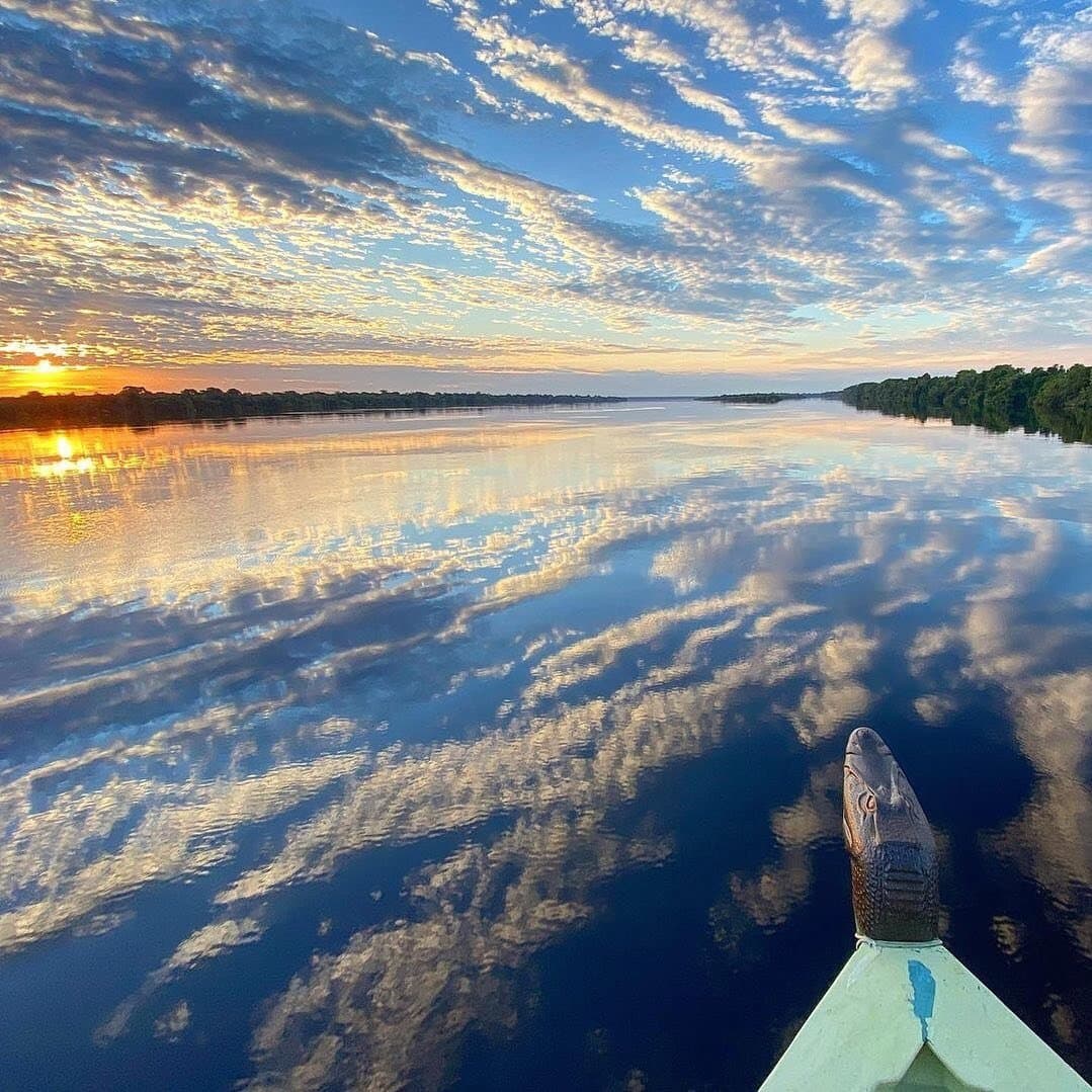 Der Fluss leiht sich den Himmel, und der Bug weiß genau, wo es langgeht. Blick vom Bug eines kleinen Flussboots auf einer Amazonas-Flusskreuzfahrt in Brasilien, mit einer geschnitzten Kaiman-Galionsfigur, die auf einen spiegelglatten Schwarzwasserfluss zeigt, der orangefarbene Sonnenaufgangswolken reflektiert, flankiert von dichtem Regenwald