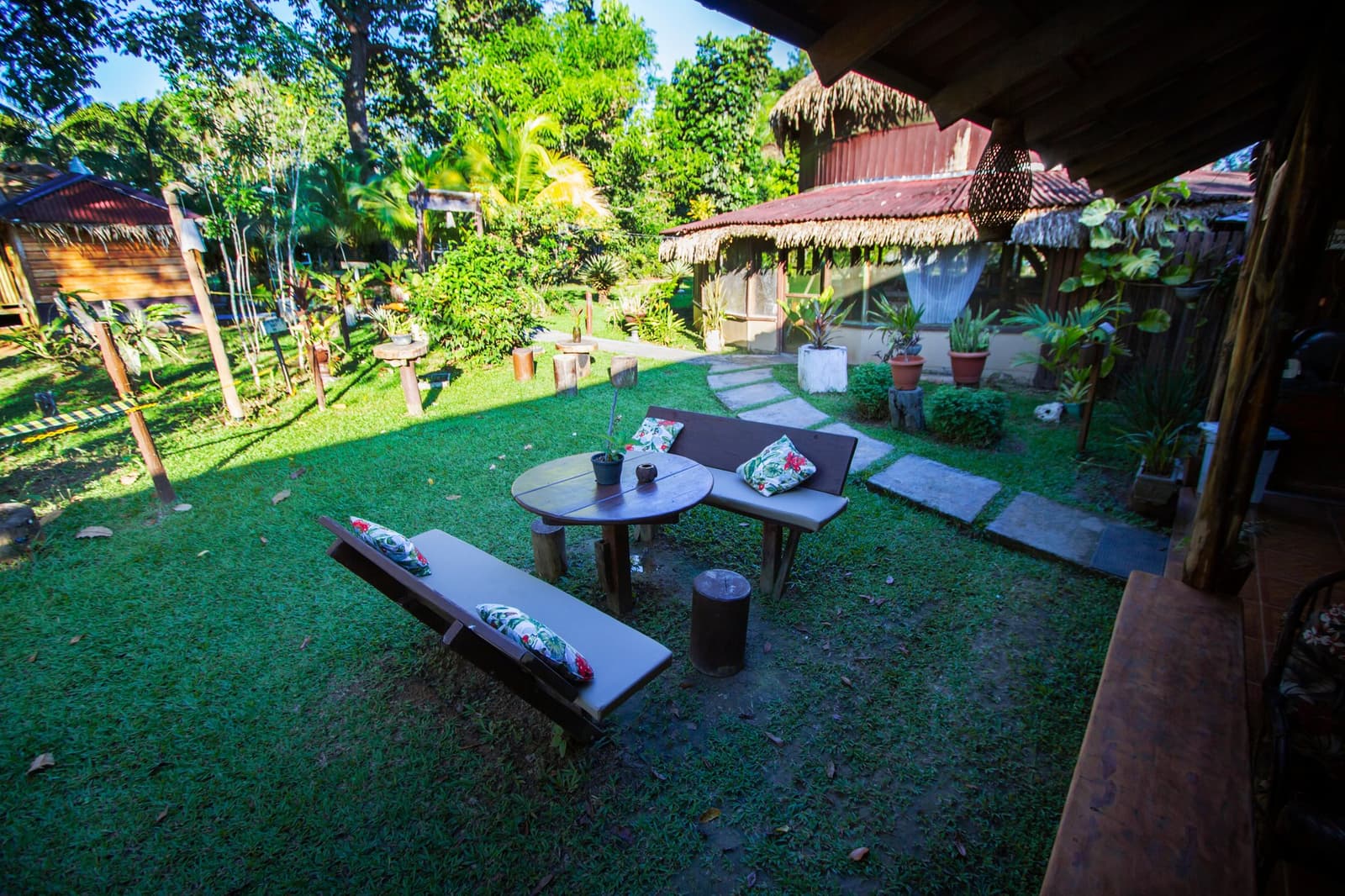Ein Garten, in dem Pläne sich still auflösen. Gepolsterte Holzbänke und ein runder Tisch auf einem gepflegten Rasen in der Amazon Turtle Lodge, mit Strohdach-Bungalows und üppigem Tropengartenblick