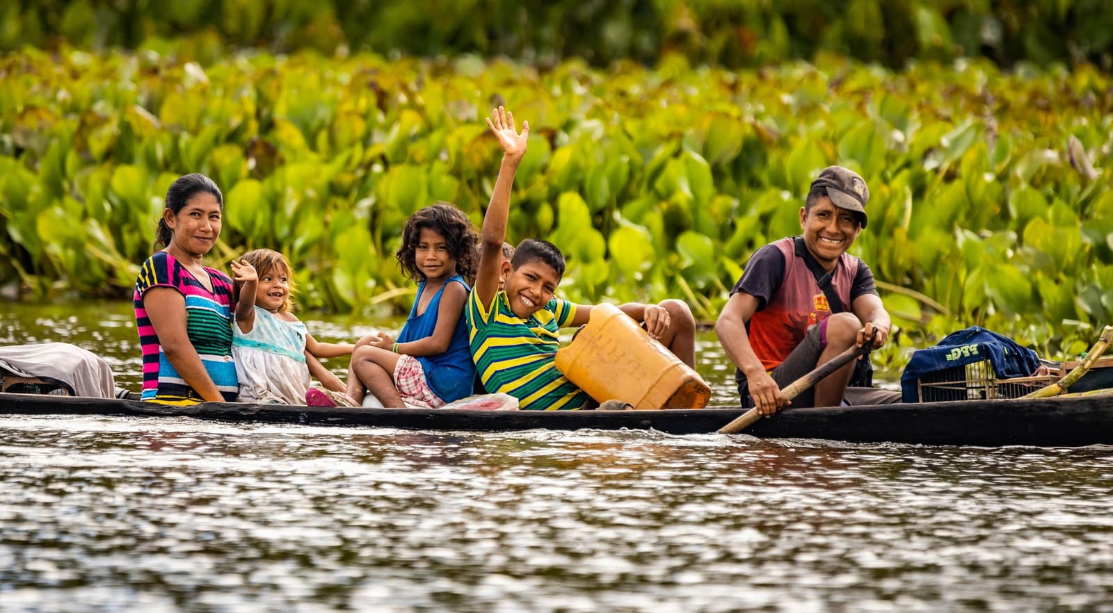 Kein Stau, kein Stress, nur Fluss und Gelächter. Eine lächelnde einheimische Familie — Mutter, Vater und drei Kinder — paddelt in einem hölzernen Einbaum an dichter grüner Wasservegetation auf einem ruhigen Amazonasfluss vorbei, wobei ein Junge fröhlich in die Kamera winkt