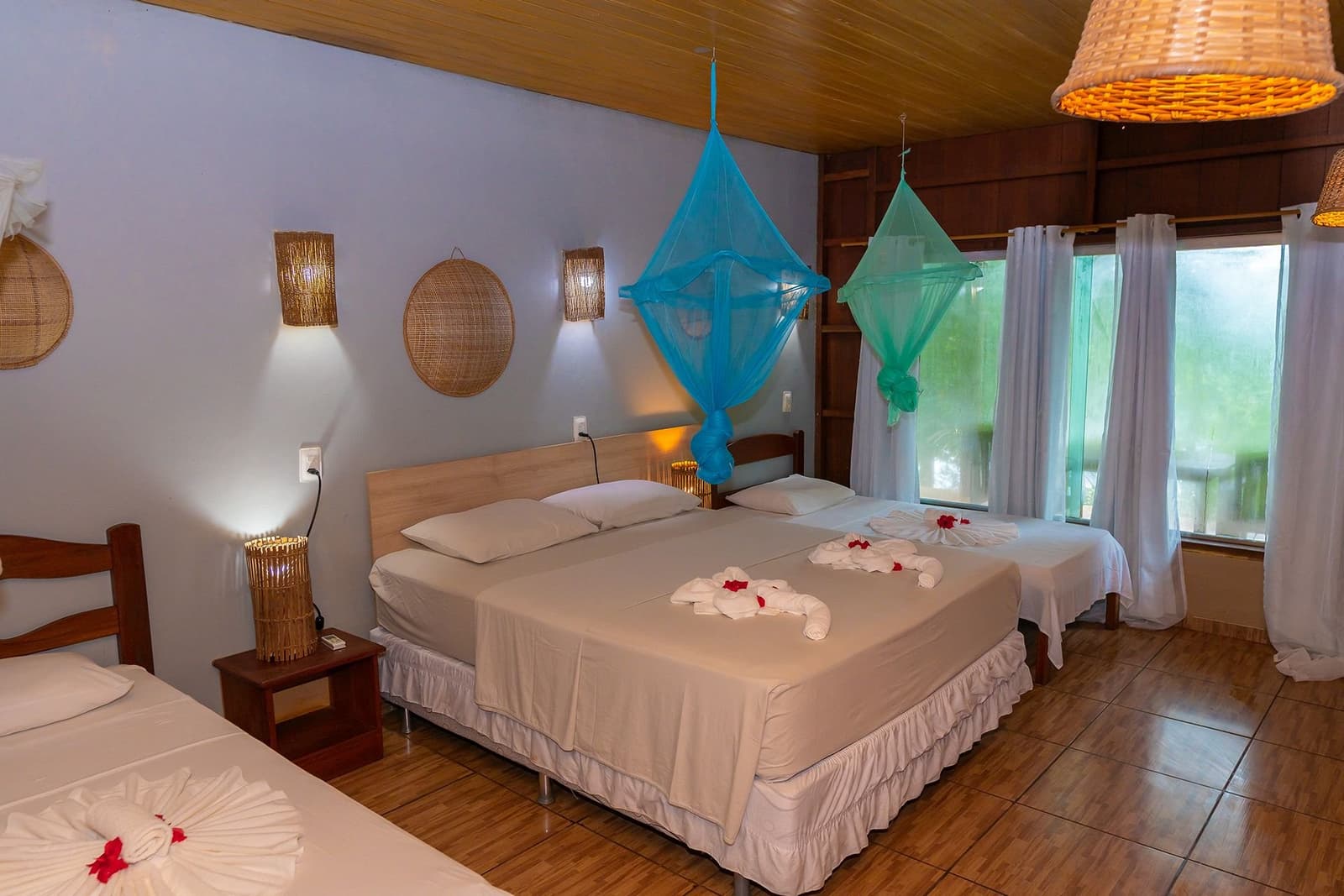 Handtuchschwäne, Betthimmel und der Regenwald direkt vor dem Fenster. Ein Dreibettzimmer in der Amazon Turtle Lodge mit farbenfrohen hängenden Moskitonetzen, gefalteten Handtuchdekorationen auf weißer Bettwäsche, gewebten Rattanlampen und einem Fenster mit Blick ins Grüne