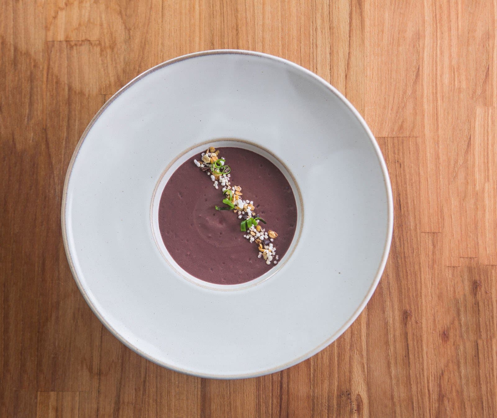 Des ingrédients sobres, une cuisine qui ne l'est pas. Méritée. Photo: Rjcastilho Vue de dessus d'une soupe crémeuse violet profond servie dans un grand bol en céramique blanche, garnie d'une ligne diagonale de graines grillées, de riz soufflé et d'oignon vert tranché, sur une surface en bois chaud dans un lodge d'observation des oiseaux en Amazonie