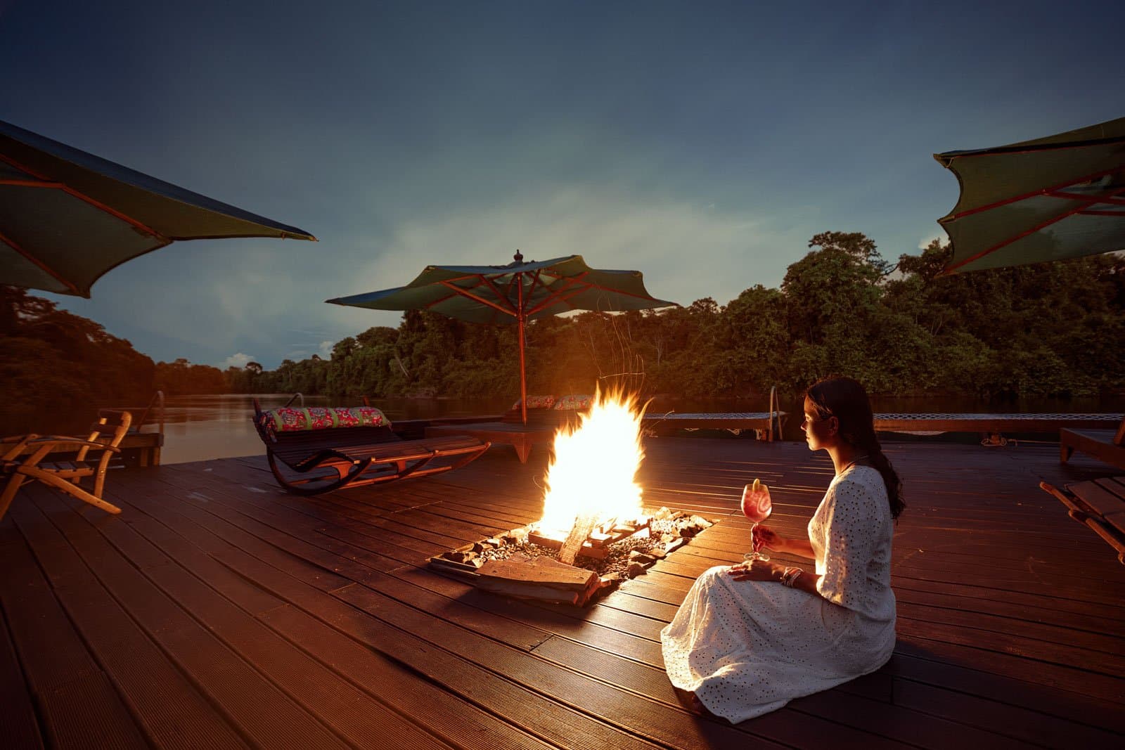 Cocktail en main, feu allumé, la forêt prend ses quartiers de nuit. Photo: Samuel Melim Une femme en robe blanche est assise près d'un feu de camp à ciel ouvert sur une terrasse en bois donnant sur une rivière forestière au crépuscule, un cocktail à la main