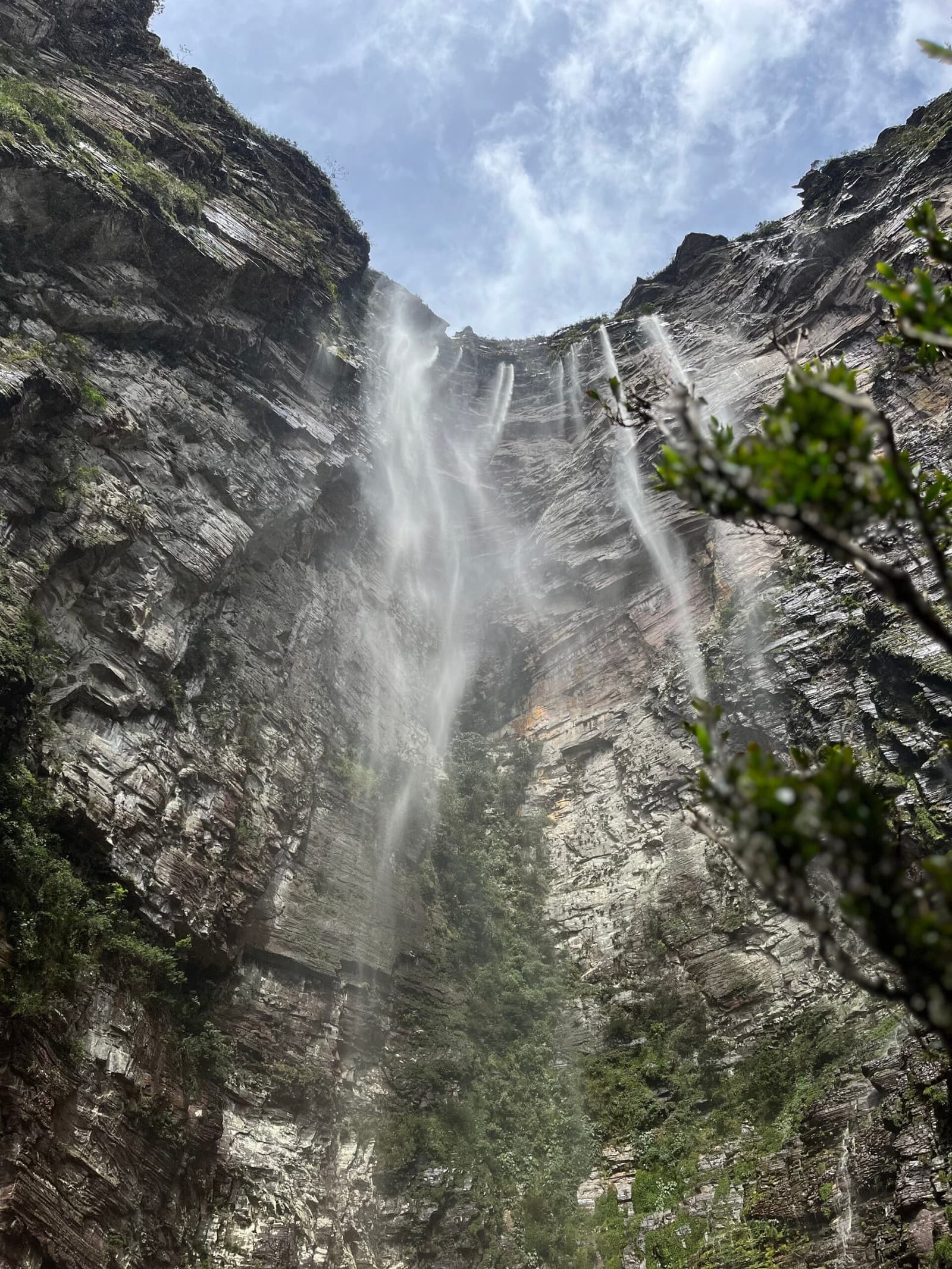 So viel Höhe, und der Nebel erreicht dich trotzdem unten. Photo: Lucas Neves Blick von unten auf den Cachoeirão-Wasserfall beim Chapada-Diamantina-Trek, mit mehreren Wassersträhnen, die eine hohe geschichtete Sandsteinfelsenwand hinabfließen, eingerahmt von grüner Vegetation und teils bewölktem Himmel