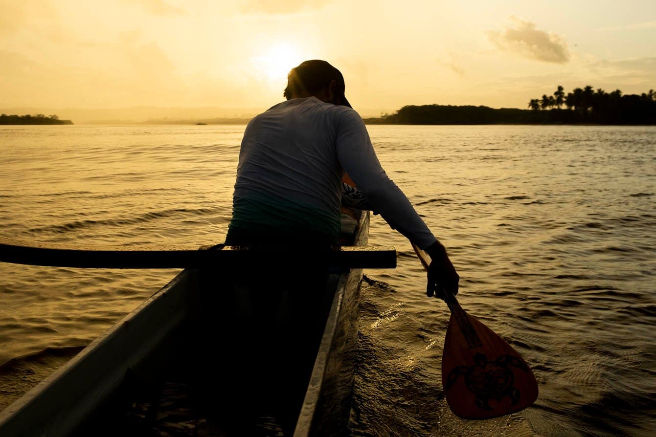 Paddel in der Hand, den Rest erledigt der Fluss. Ein Paddler im hawaiianischen Kanu taucht sein Holzpaddel in ruhiges Wasser, als Silhouette vor einem goldenen Abendhimmel mit bewaldeter Küste im Hintergrund