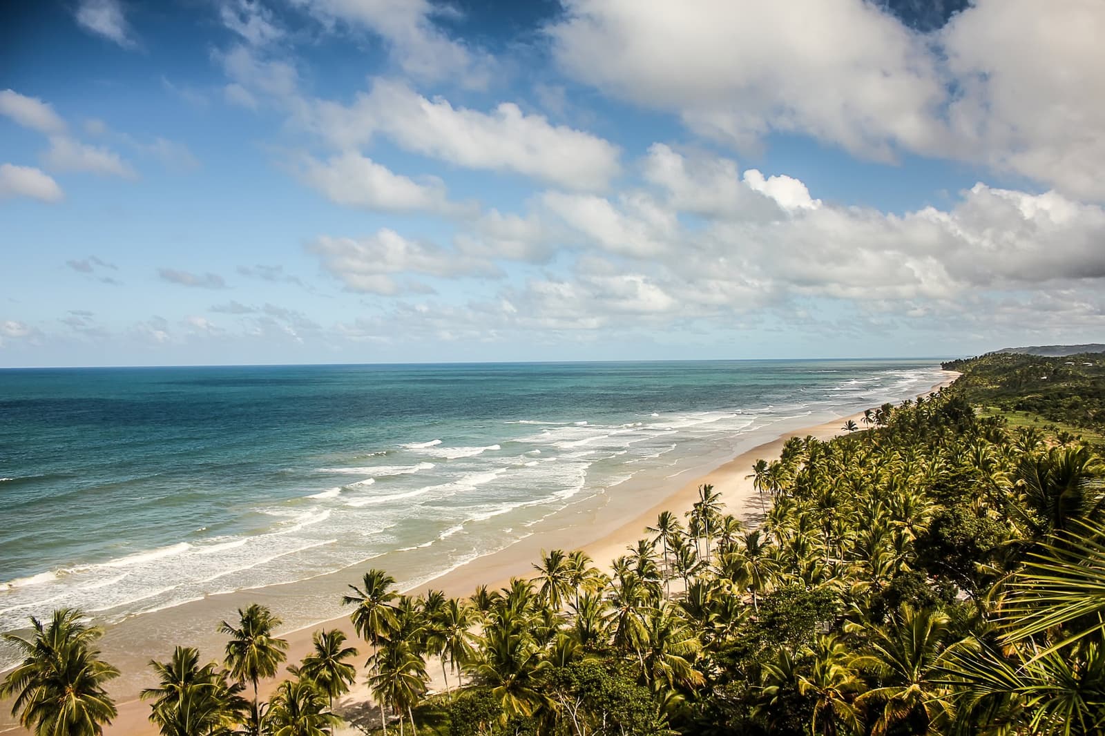 Kilometerlanger Sand, kaum eine Menschenseele, und die Palmen stehen Spalier. Photo: MaFelipe Luftaufnahme eines langen Sandstrandes, gesäumt von dichten Kokospalmen und Atlantischem Regenwald, mit türkisfarbenen Wellen unter einem teilweise bewölkten Himmel auf der Bahia Radroute