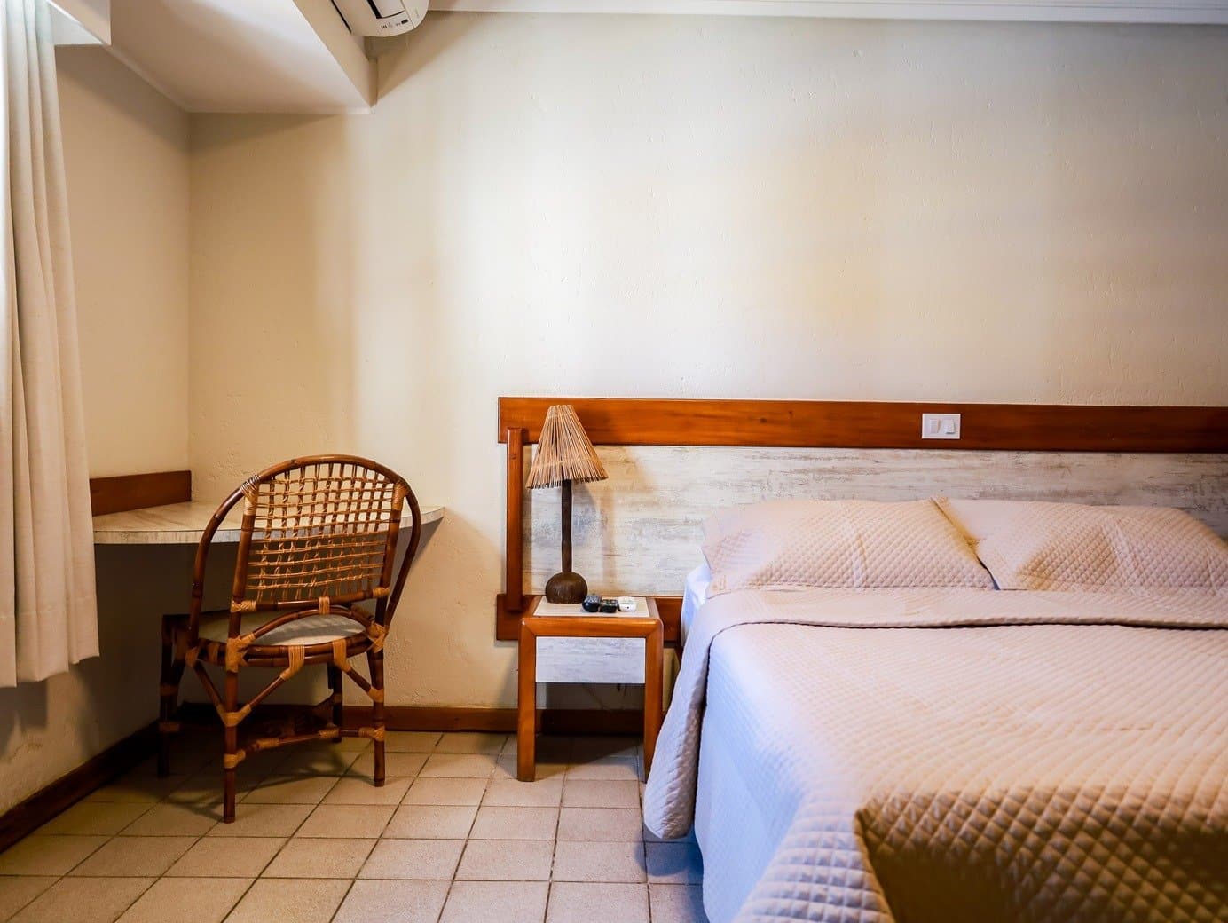 Schlicht, sauber und genau das, was müde Beine brauchen. Einfaches, gemütliches Gästehauaszimmer auf einer Bahia-Radtour mit Doppelbett, hellem Steppbettbezug, Rattanstuhl, Holzkopfteil und einer geflochtenen Bambustischlampe