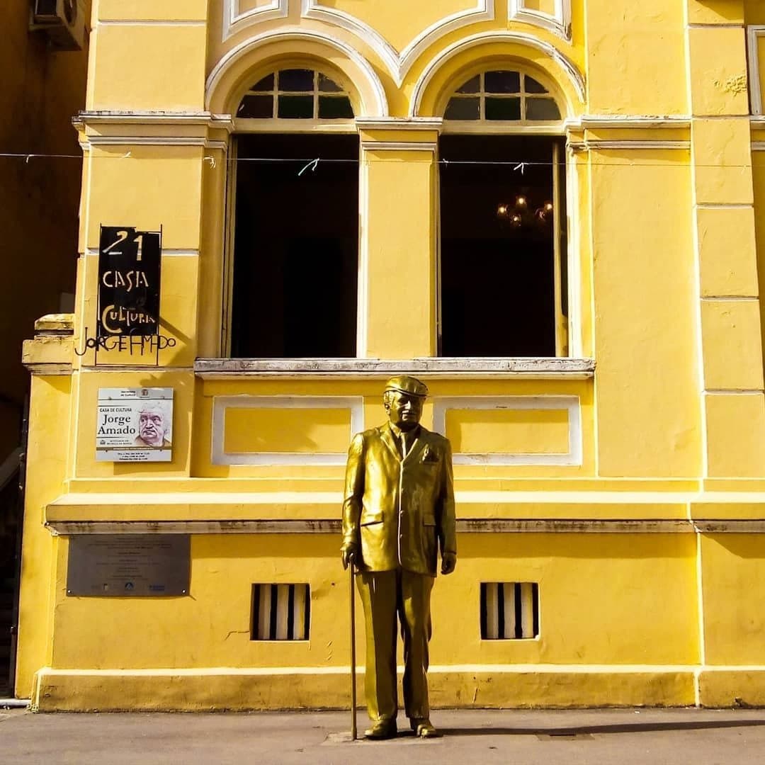 Er hat die ganze Region in eine Legende verwandelt. Hier beginnt die Geschichte. Goldene Statue von Jorge Amado mit Gehstock vor der leuchtend gelben Fassade des Casa de Cultura Jorge Amado in Ilhéus, Bahia