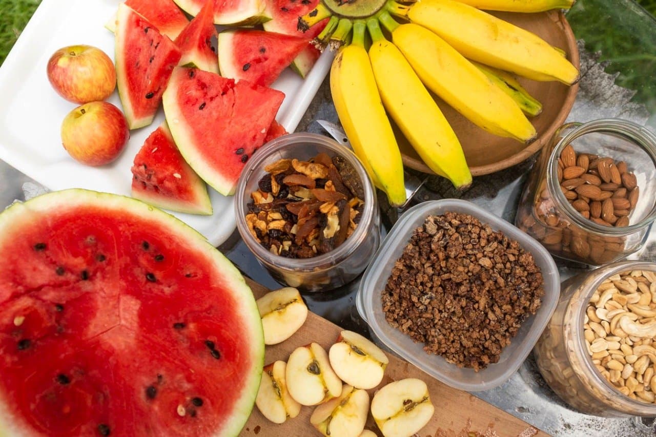 Wassermelone, Nüsse, Granola, Energie für die nächste Etappe. Vogelperspektive auf Snacks für eine Bahia-Radtour: geschnittene Wassermelone, Bananen, Äpfel, Granola, getrocknete Früchte, Mandeln und Cashews auf einem Tablett im Freien