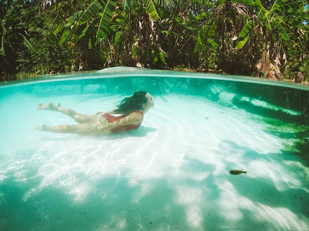 Água tão limpa que dá pra ver o peixinho curioso lá embaixo. Foto meio submersa de mulher de biquíni vermelho flutuando em um fervedouro de águas cristalinas cercado de vegetação tropical densa em uma aventura no Jalapão